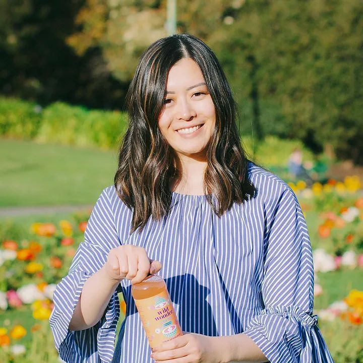 Woman in blue striped shirt smiles, holding a beverage bottle outdoors in a park with flowers and greenery.
