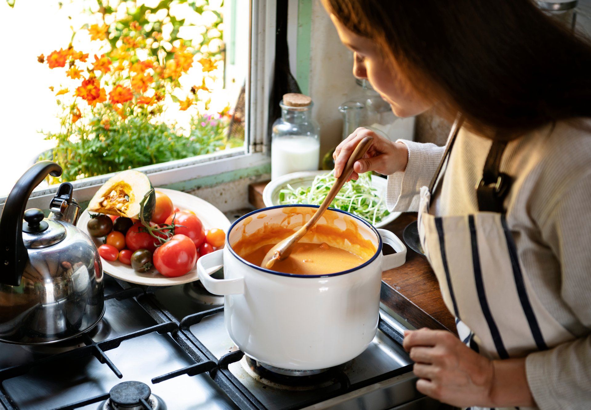 Woman stirring soup in a pot on a stovetop, window with garden view, tomatoes and squash on counter.