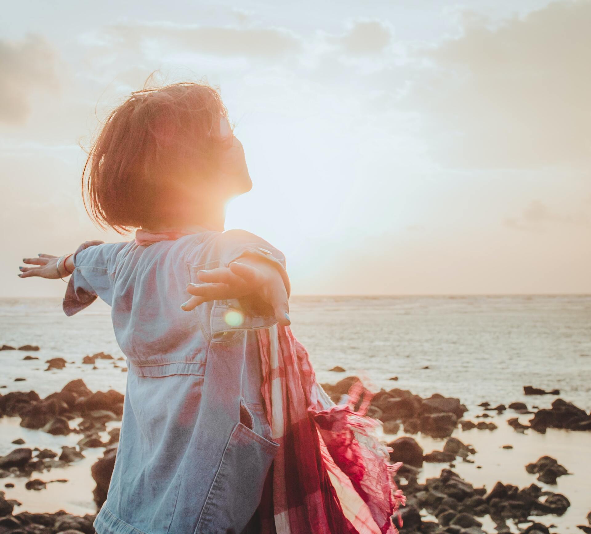 Woman with arms outstretched stands on a rocky shore, facing the setting sun over the ocean.