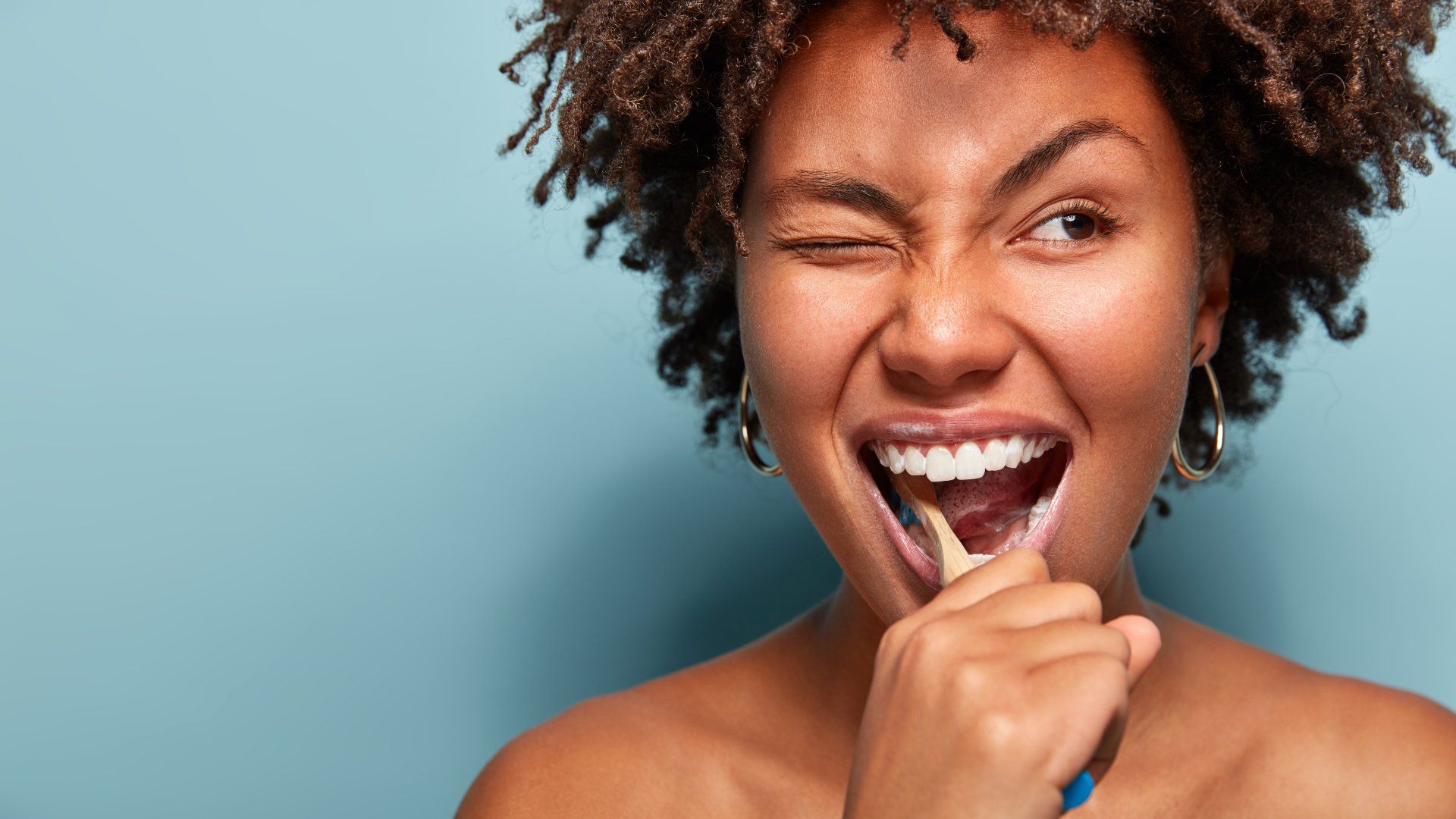 Woman brushing teeth, winking with a big smile, against a light blue background.