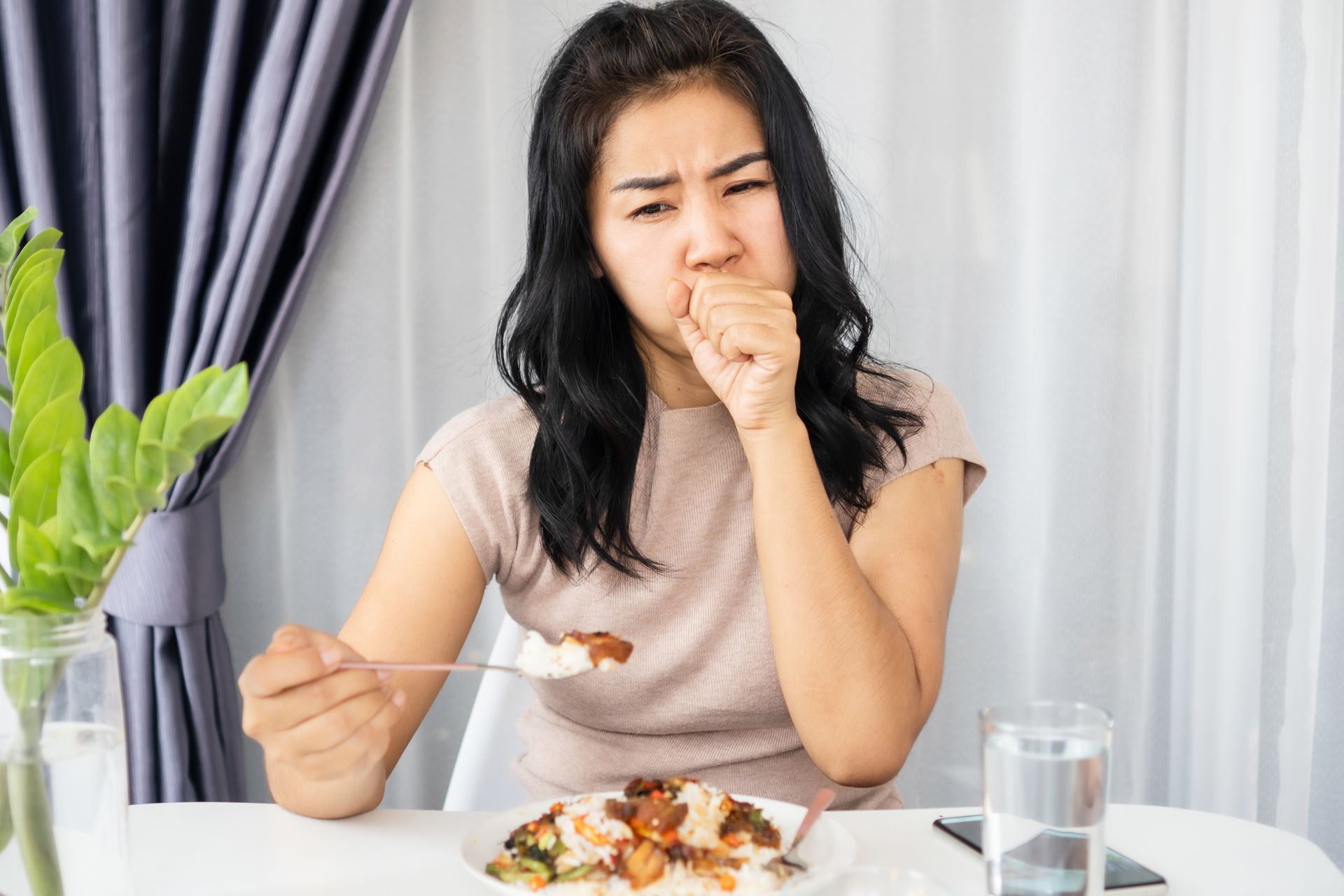 Woman coughing while eating at a table, looking displeased. Plate of food and glass of water present.