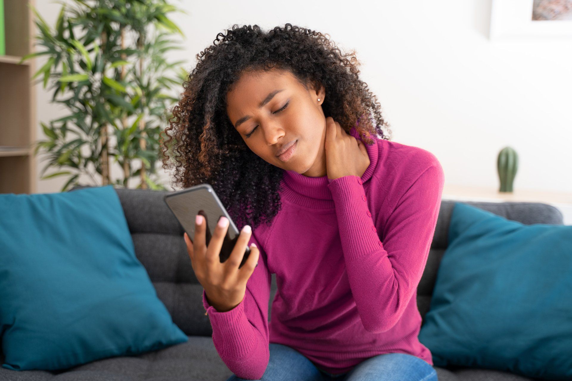 Woman with curly hair holding a smartphone, touching her neck, appearing to be in pain while sitting on a couch indoors.