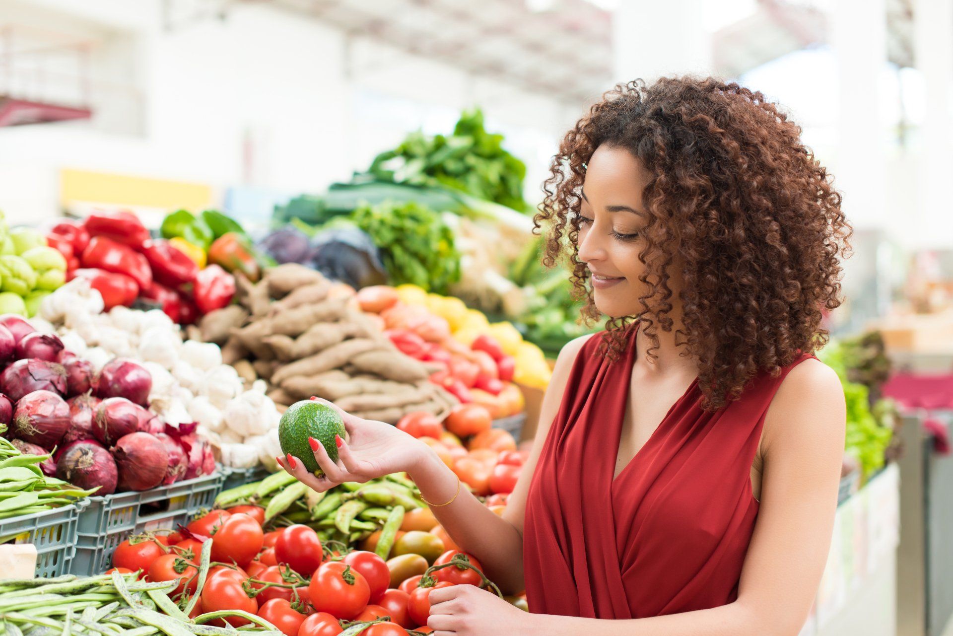A woman with curly hair in a red dress examines an avocado at a colorful produce market.