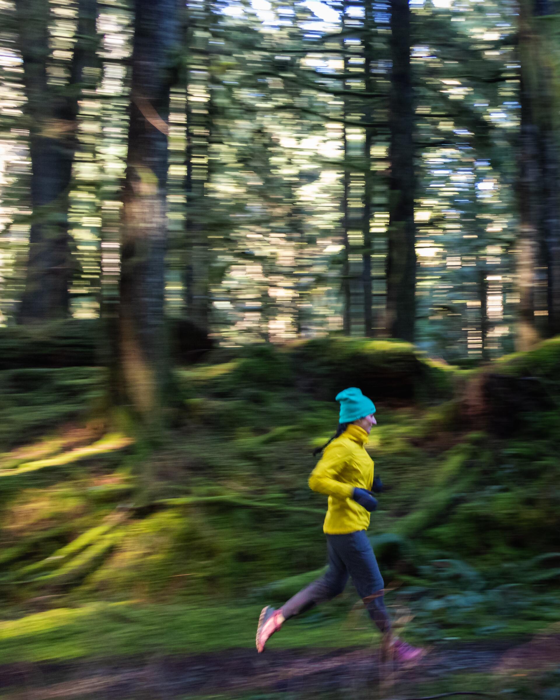 Woman jogging in a forest, wearing a yellow jacket and blue hat. Trees are blurred with motion.