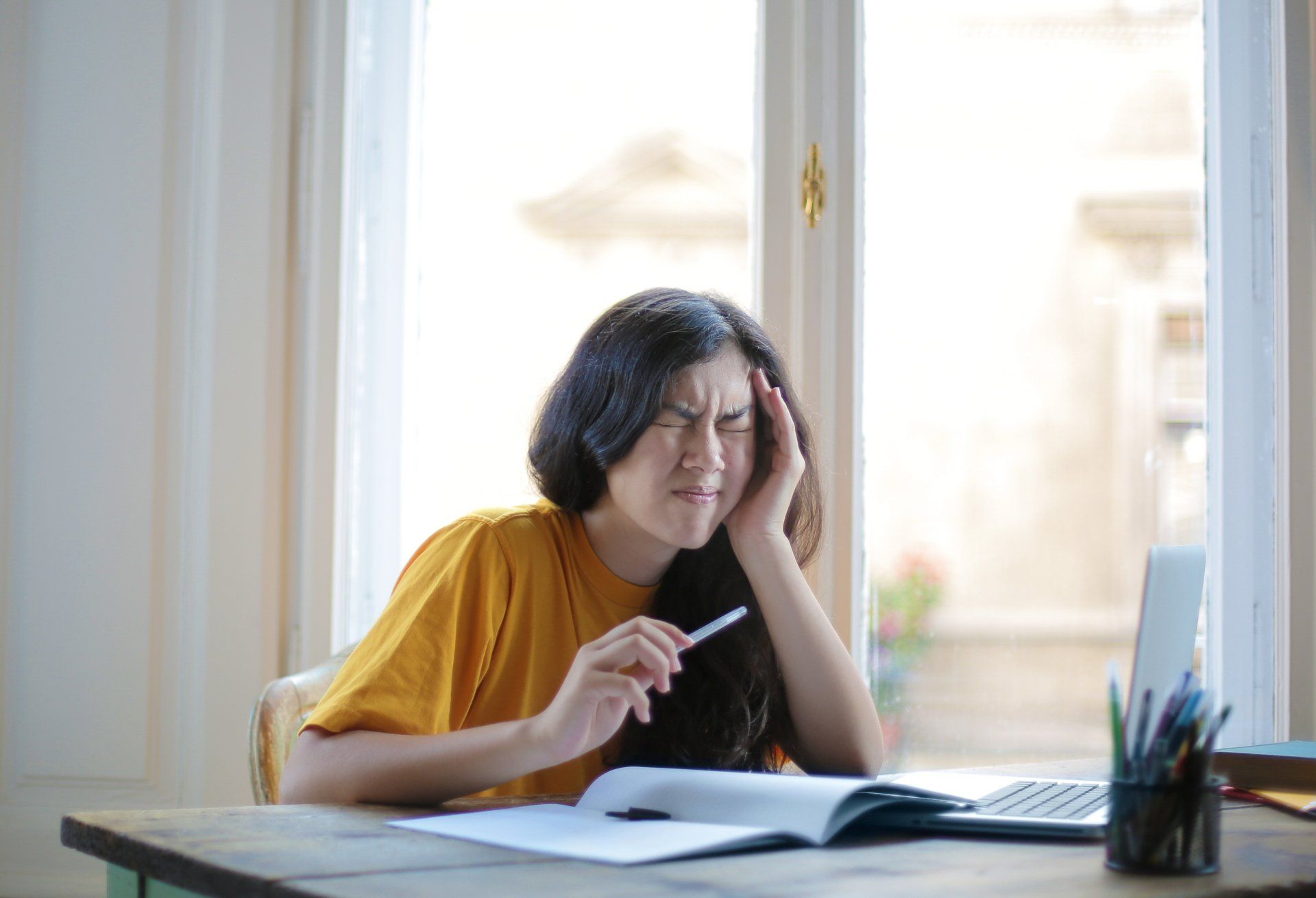Woman with dark hair in a yellow shirt, looking stressed while holding a pen. Sitting at a desk with a laptop and notebook.