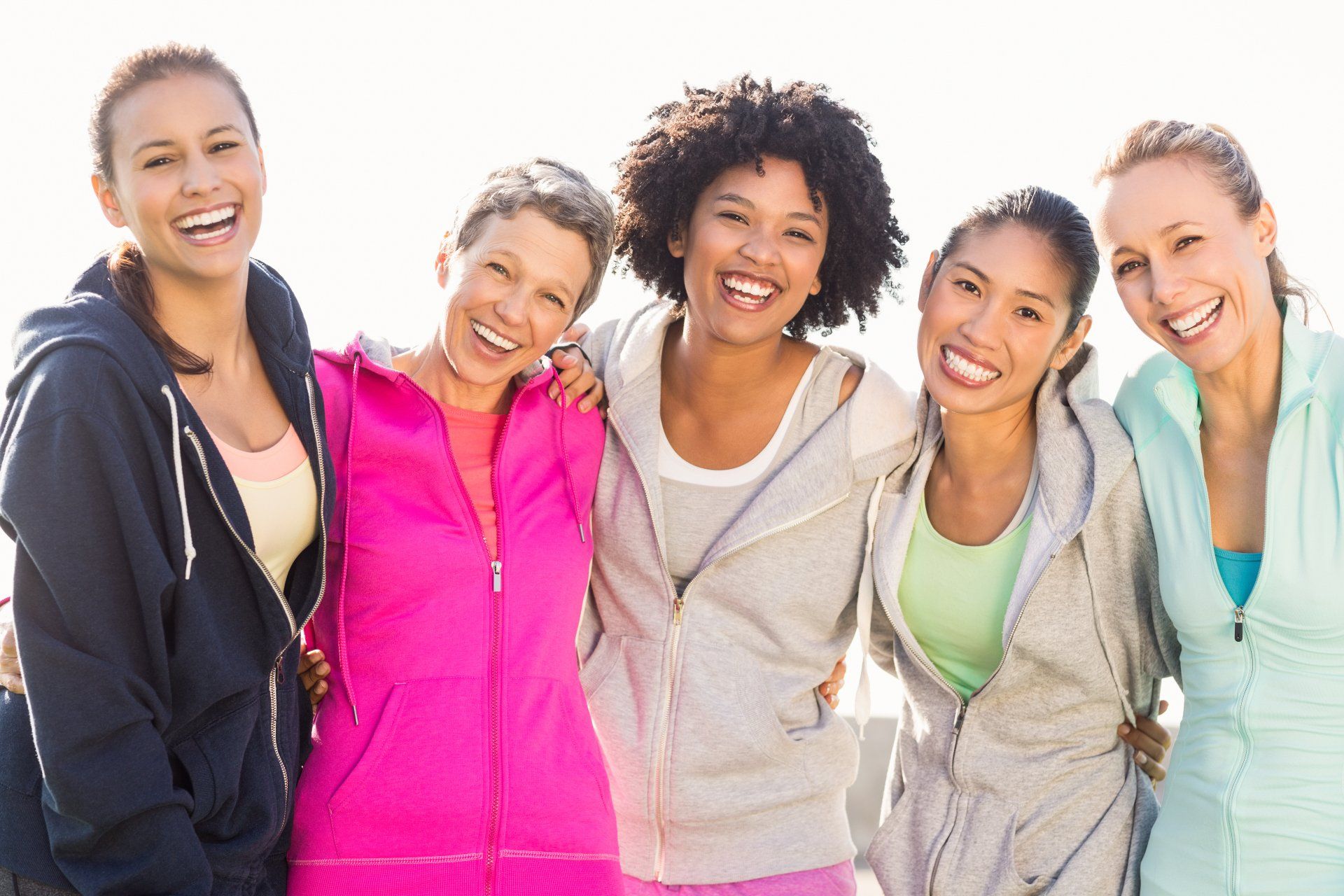 Five diverse women in workout clothes smile and hug each other outdoors.