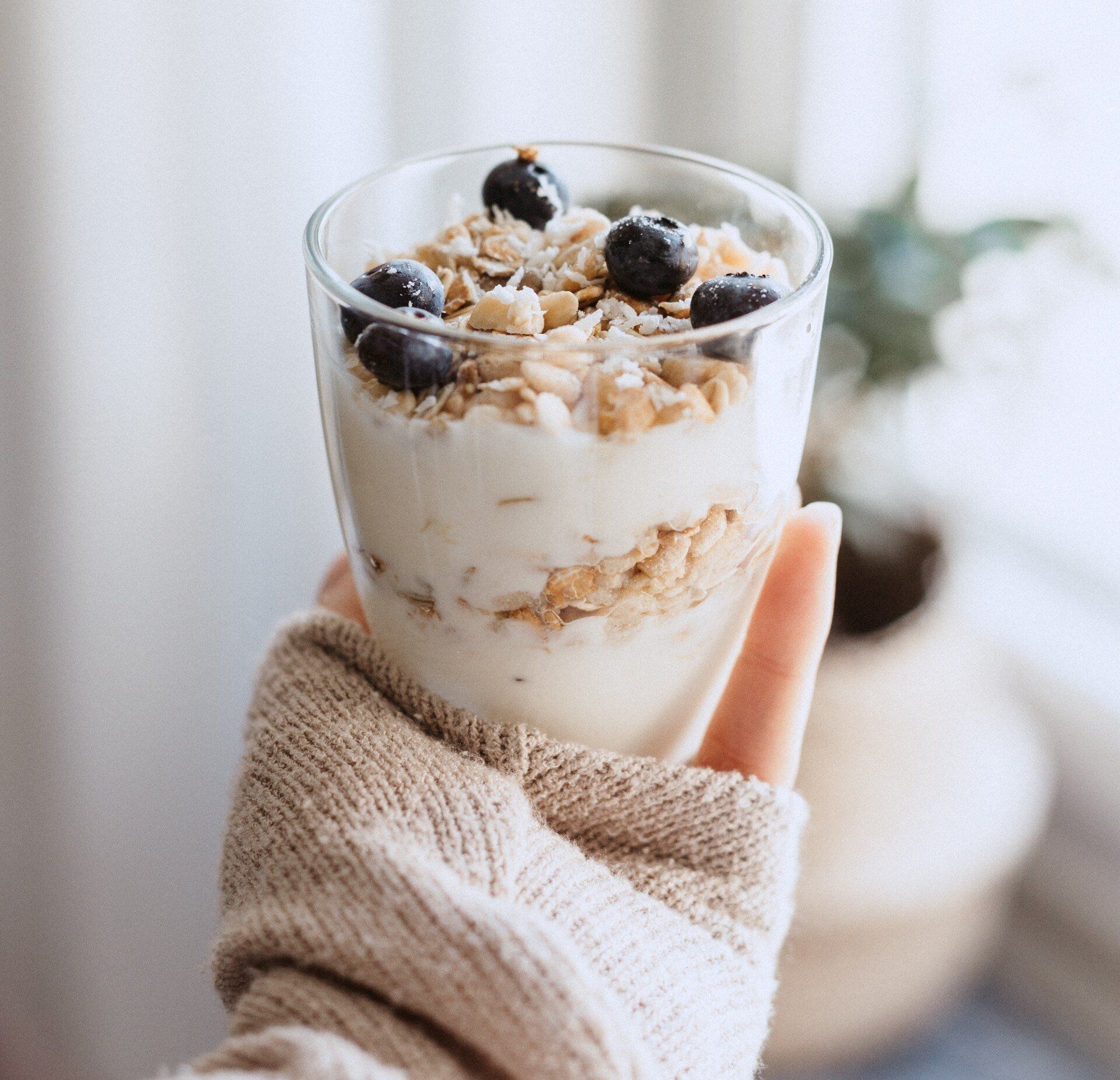 Hand holding a clear glass of layered yogurt, granola, and blueberries. The hand wears a tan sweater; blurred background.