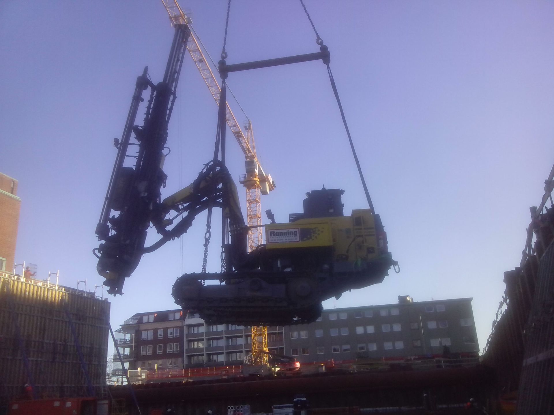 Kran løfter tungt anleggsutstyr på en byggeplass mot en blå himmel.