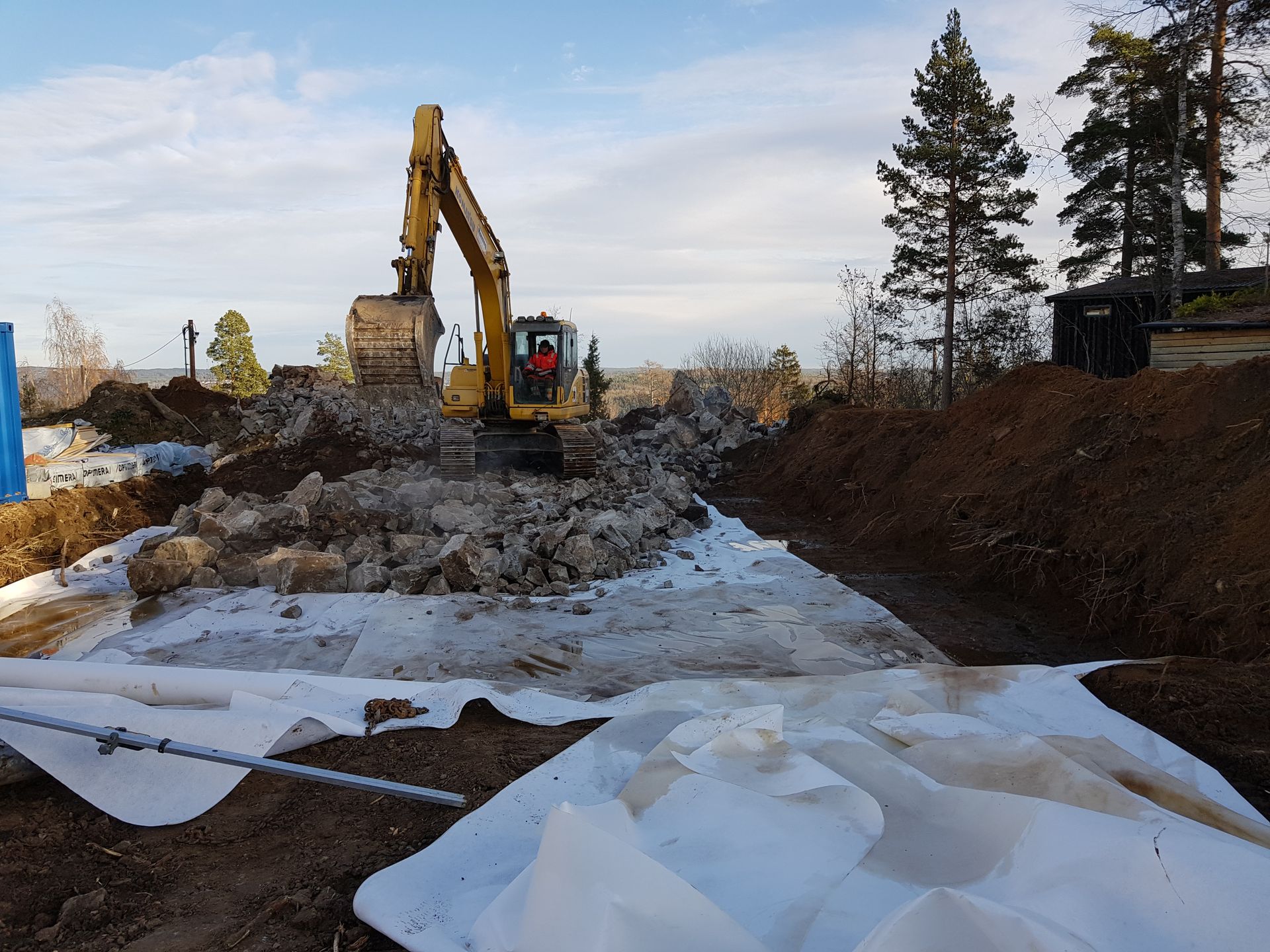 Gravemaskin fjerner rusk og avfall på en byggeplass; hvitt stoff lagt ut; trær i bakgrunnen.