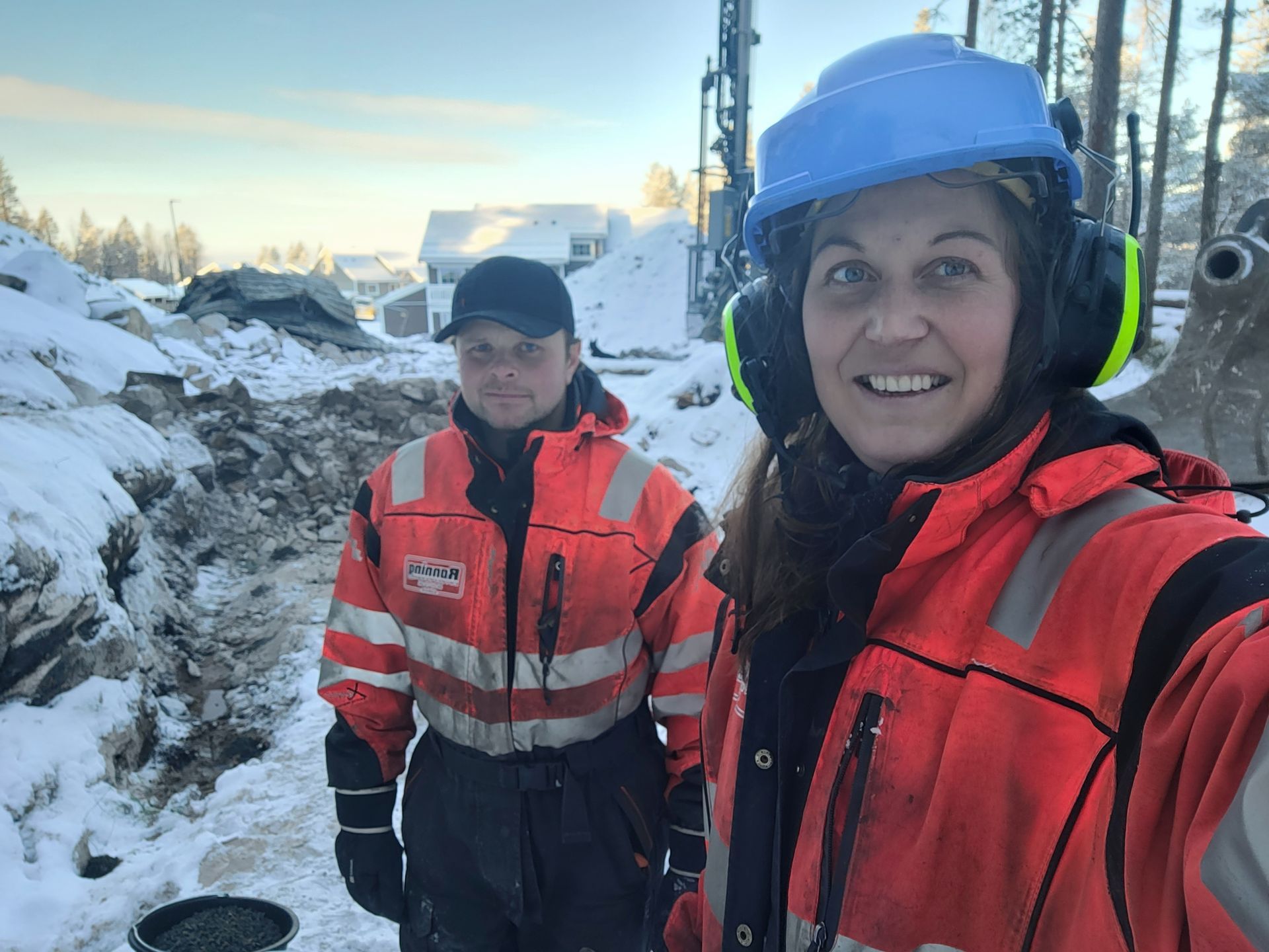 To personer i oransje arbeidsjakker poserer ved en snødekt byggeplass. Den ene bruker hjelm og hørselsvern.