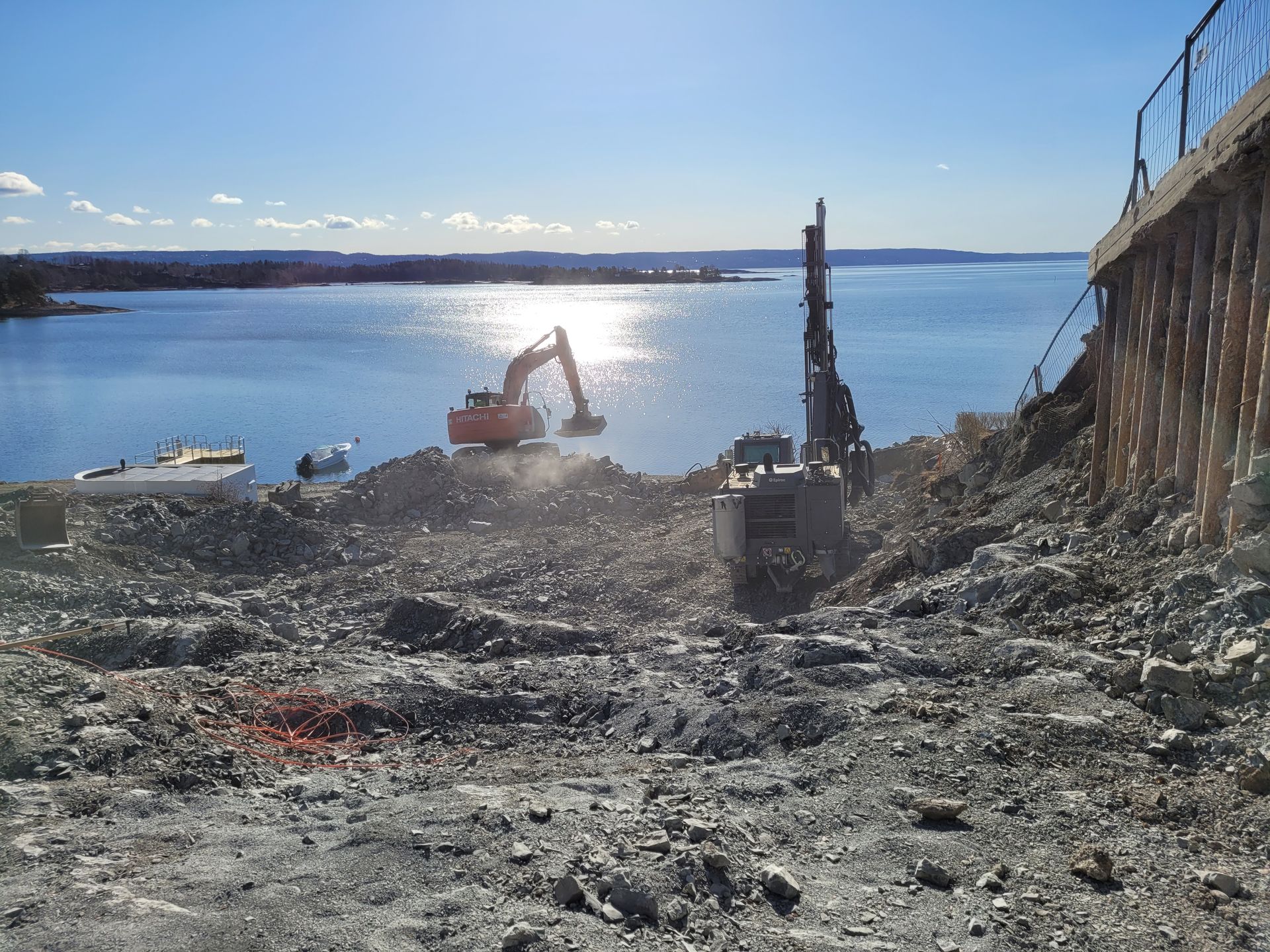 Byggeplass ved vann med gravemaskiner, steiner og en sterk sol som speiler seg i vannet.