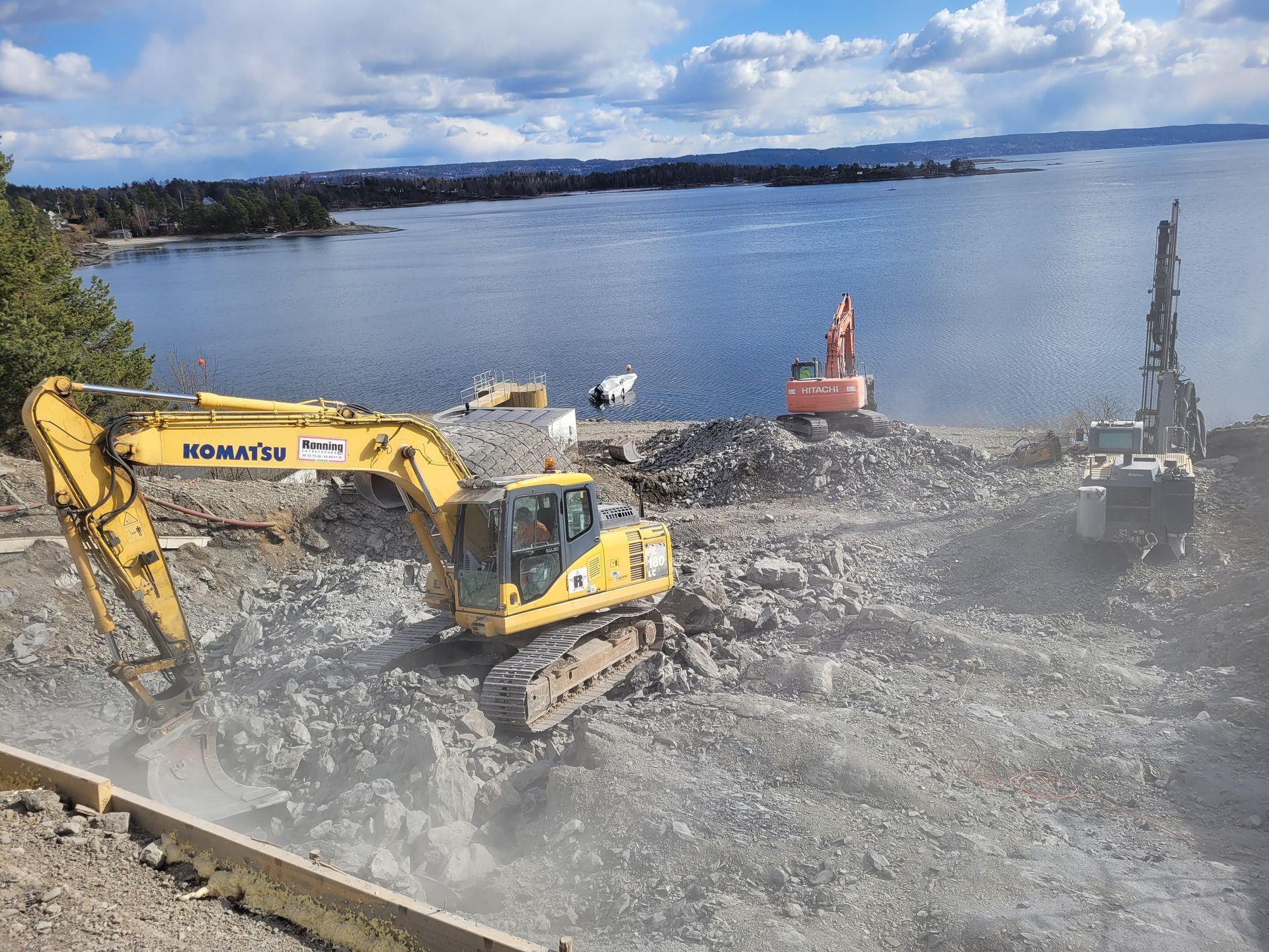 Gravemaskiner jobber på en steinete strand, nær et vann. Blå himmel i bakgrunnen.
