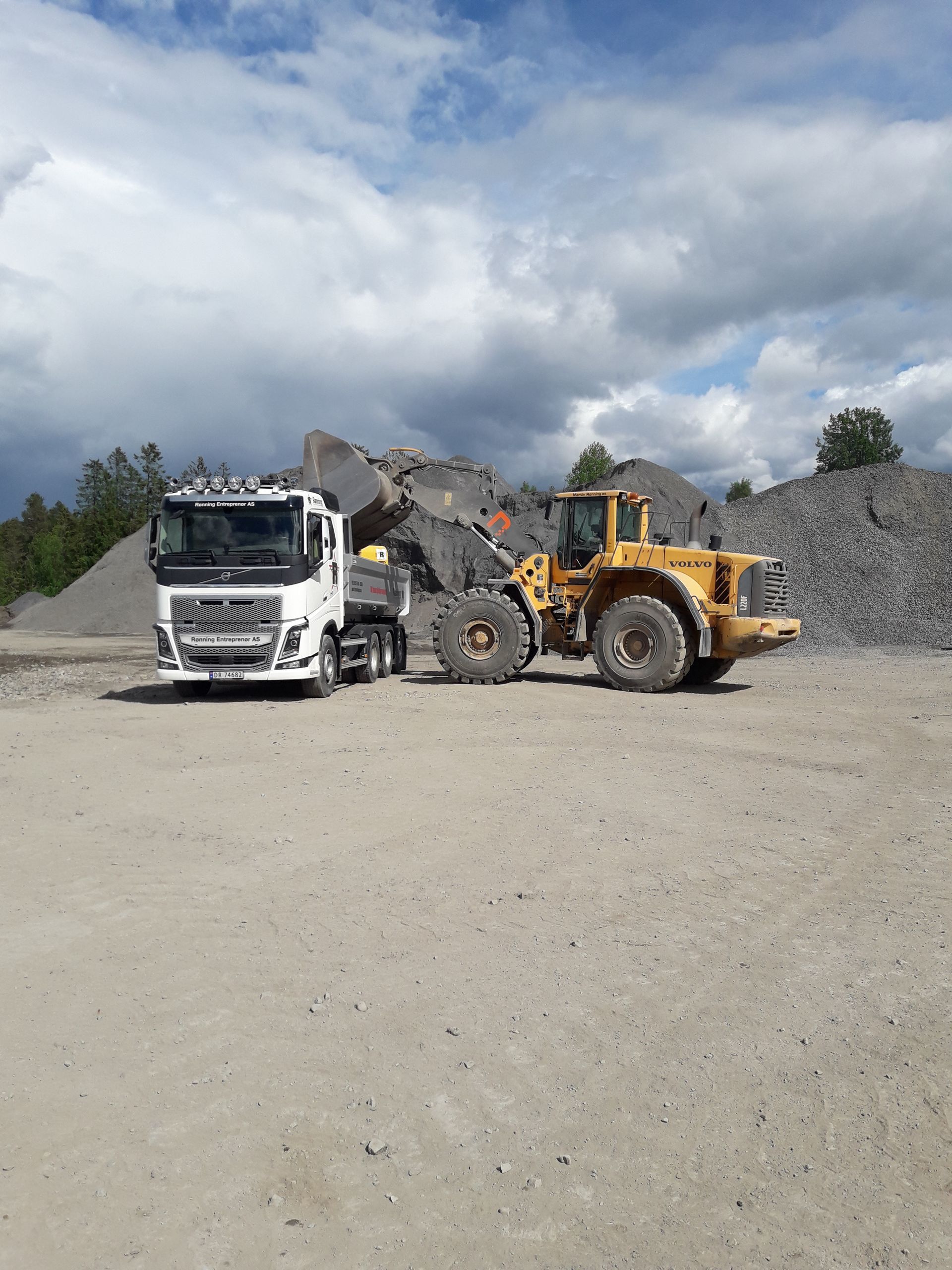 En gul laster fyller en hvit dumper med grå grus ved en grustak under en overskyet himmel.