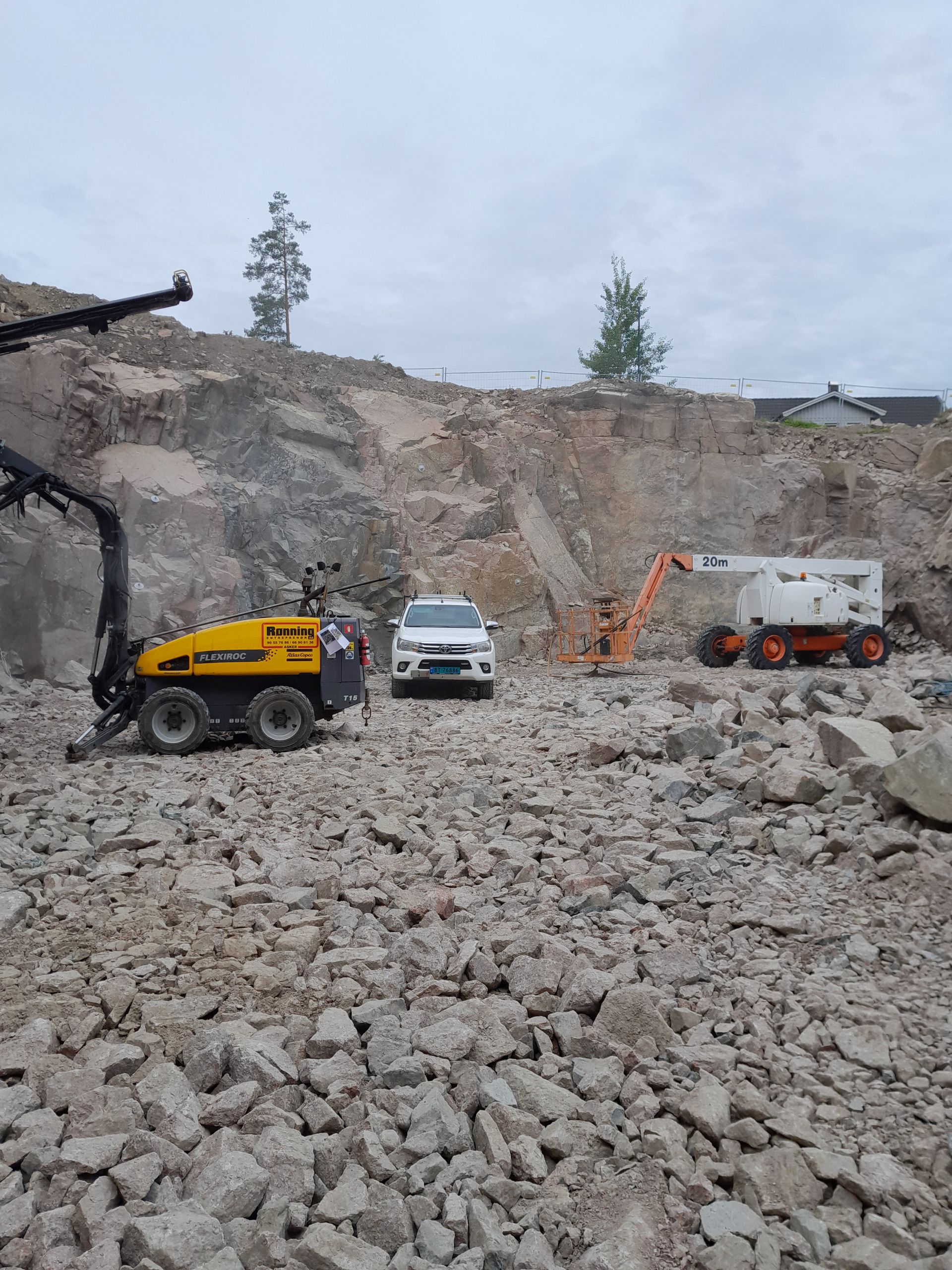 Byggeplass: hvitt kjøretøy, gult maskineri og oransje utstyr blant steiner og en steinbruddsmur. Overskyet himmel.