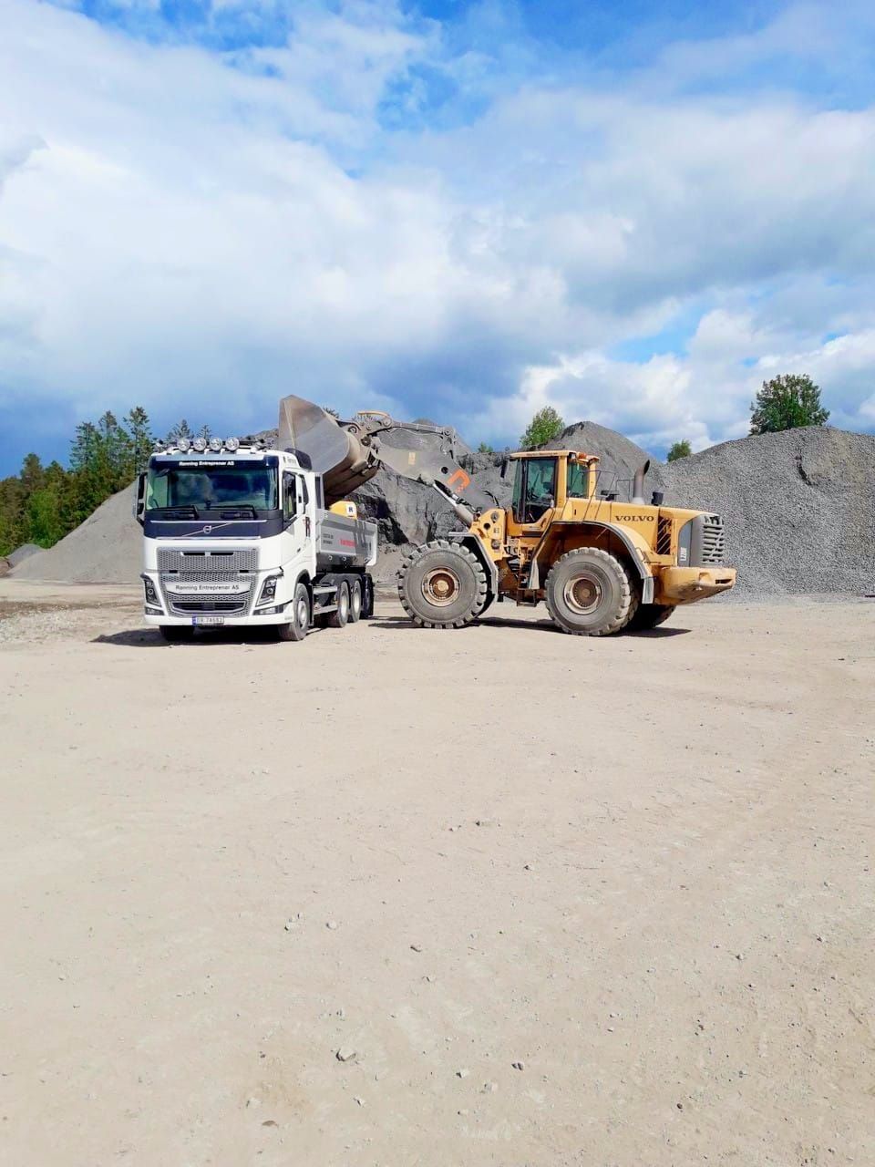Gul laster fyller en hvit lastebil med grus ved et steinbrudd under en overskyet himmel.