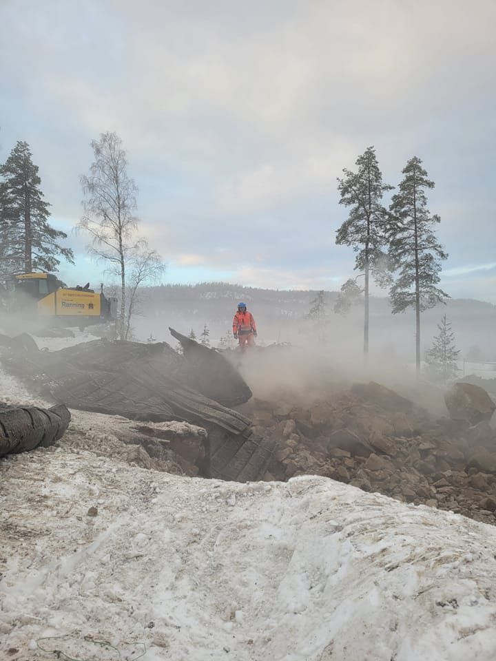 En person i oransje jakke står midt i et røykfylt landskap med snø, trær og tungt maskineri i bakgrunnen.