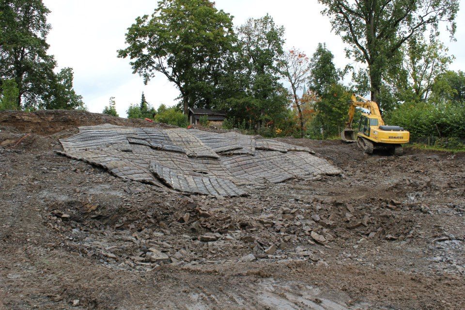 En gravemaskin og trematter i en åsside, muligens for stabilisering, med trær i bakgrunnen.