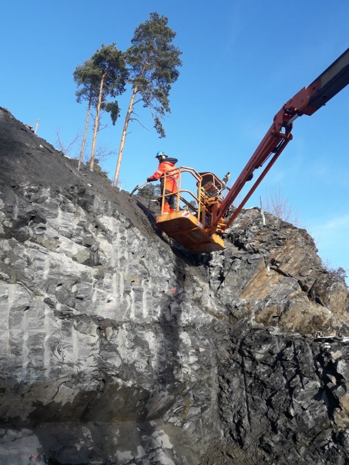 Person i en oransje heisplattform som arbeider på en steinete klippevegg, trær i bakgrunnen, blå himmel.