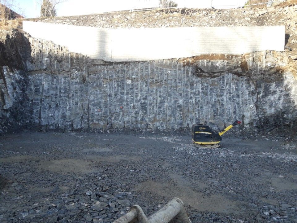 Byggeplass med en steinmur skåret med vertikale spor; en komprimator står ved foten.