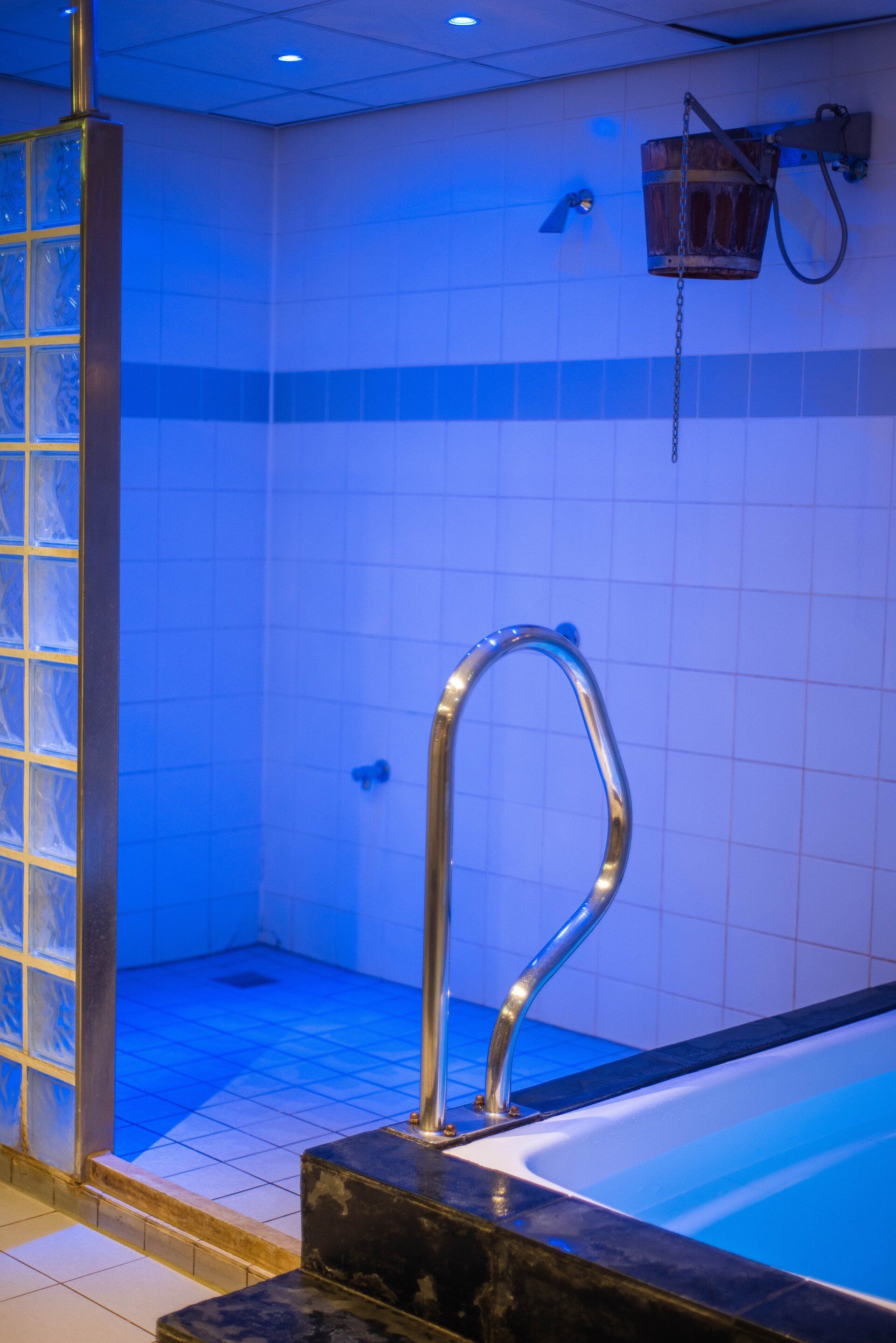Blauw verlichte spa-doucheruimte met een klein zwembad, een wand van glazen blokken en een houten emmer.