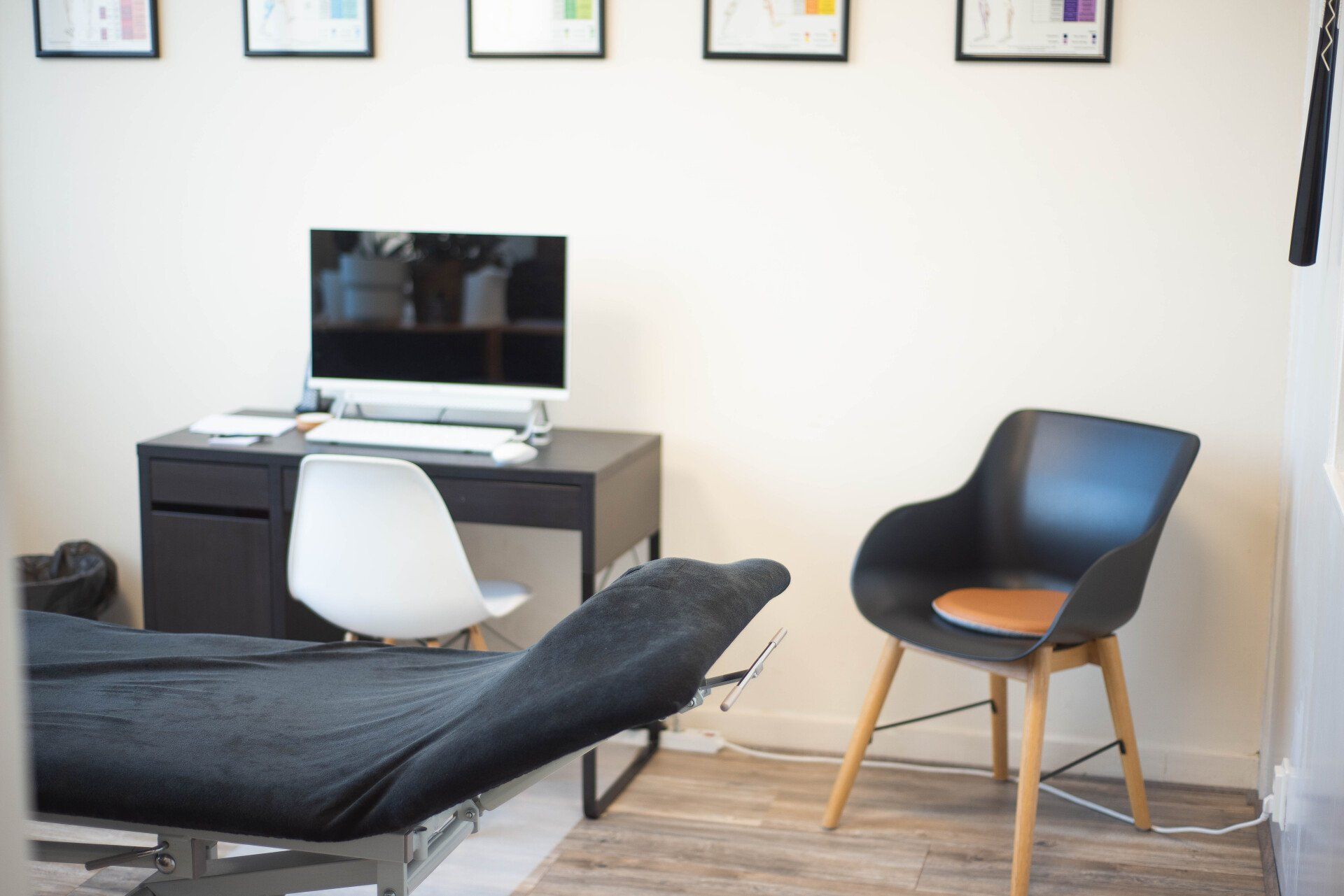 Therapieruimte: zwart medisch bed, bureau met computer, zwart-witte stoelen, witte muren.