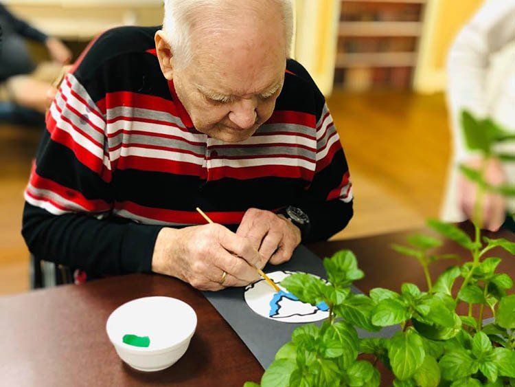 An elderly man is sitting at a table painting with a brush.