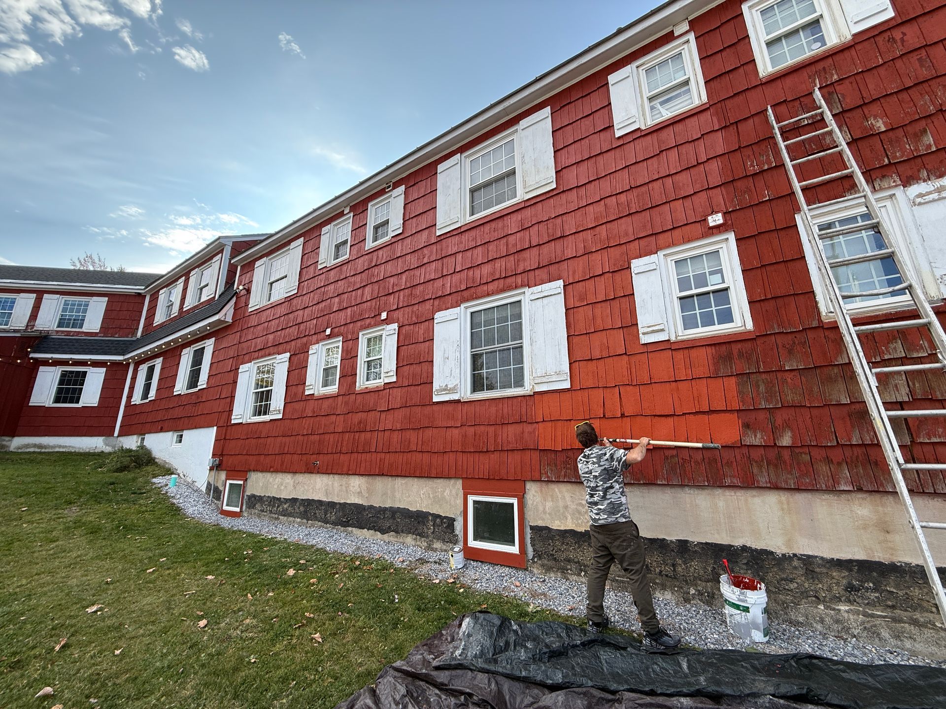 Person painting the red exterior of a building with white shutters; ladder propped against the side.