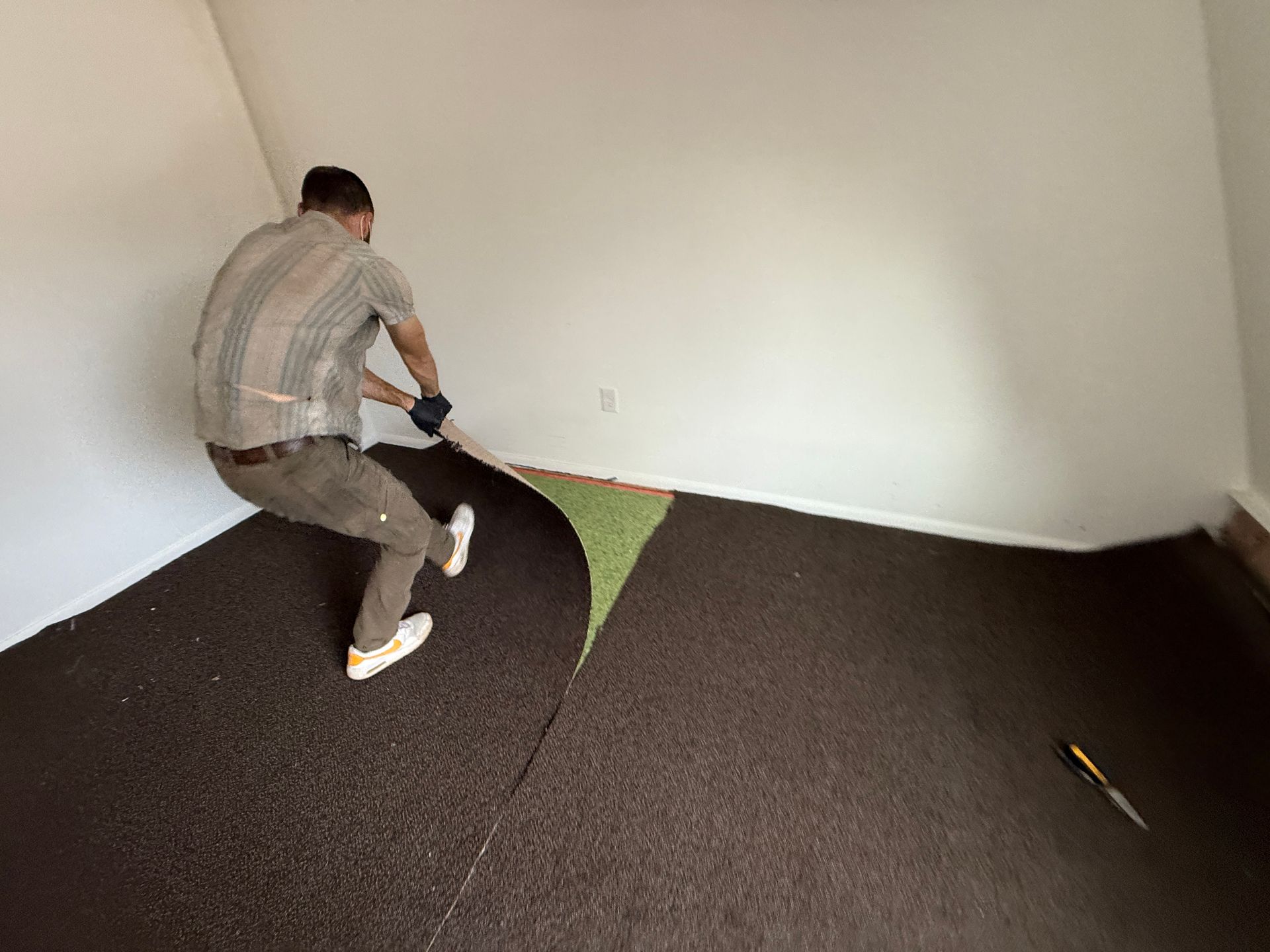Man pulling up brown flooring in a room with white walls, a utility knife on the floor.