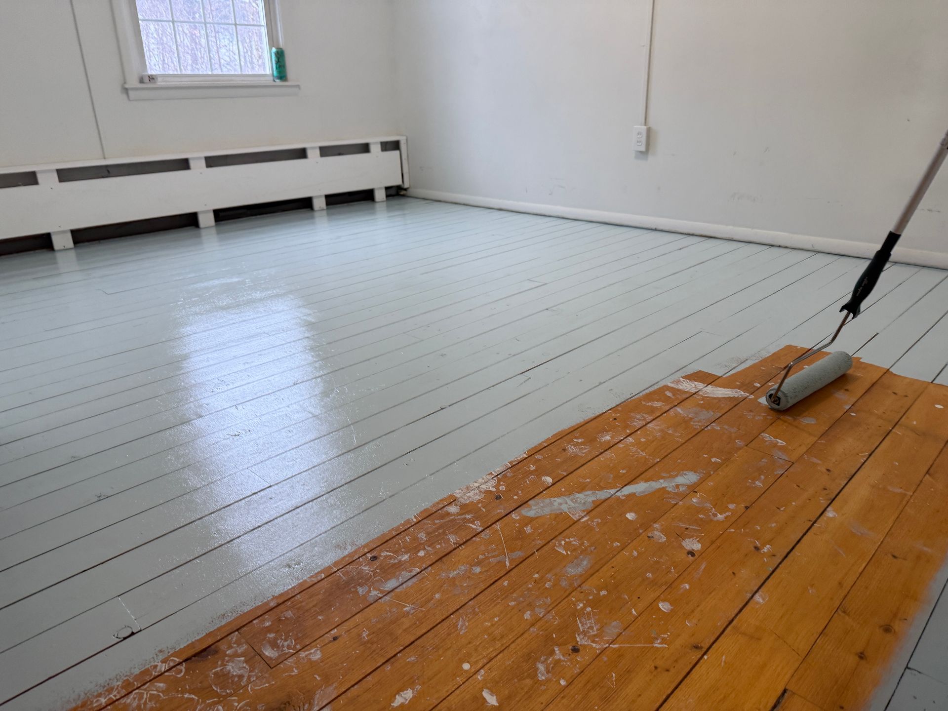 Wooden floor being painted light blue with a roller. White walls, radiator, and window visible.