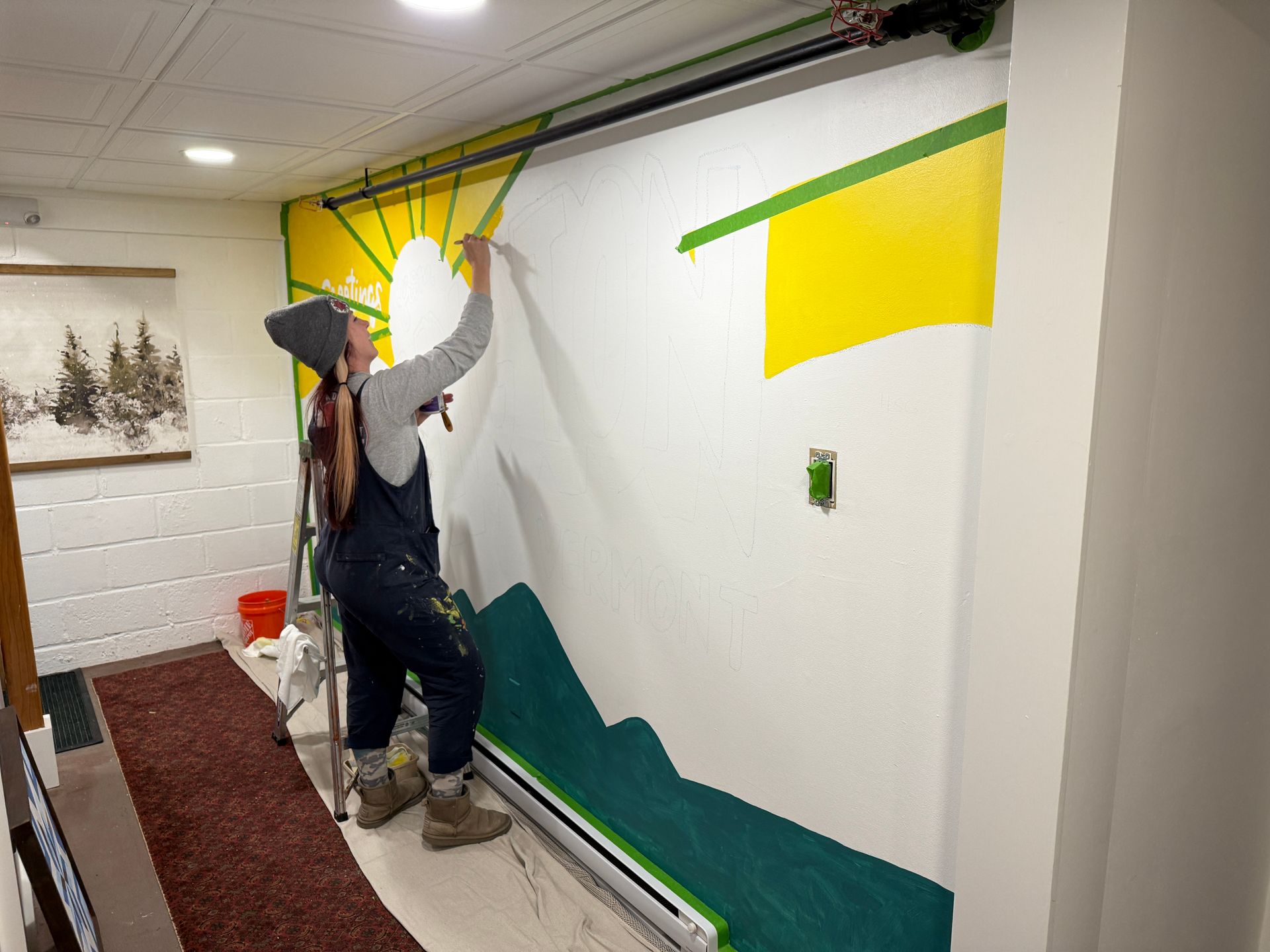 Person painting a mural of a mountain landscape with a yellow sun.