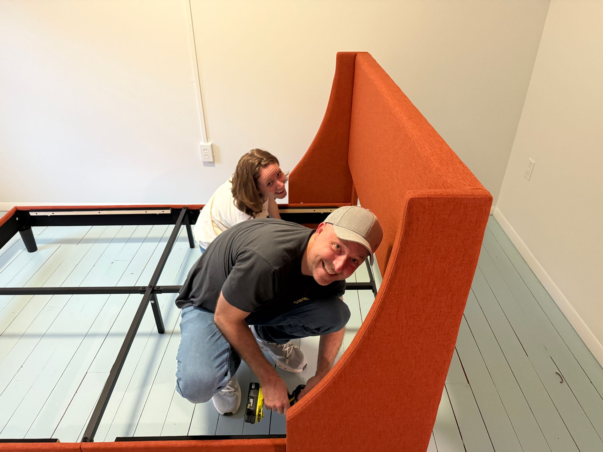 Two people assembling an orange upholstered bed frame in a room with light blue floors and white walls.