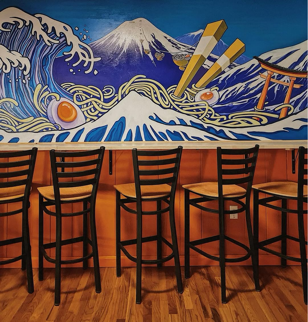 Bar seating with ramen mural of Mt. Fuji and wave in background. Orange wall, wooden floors.