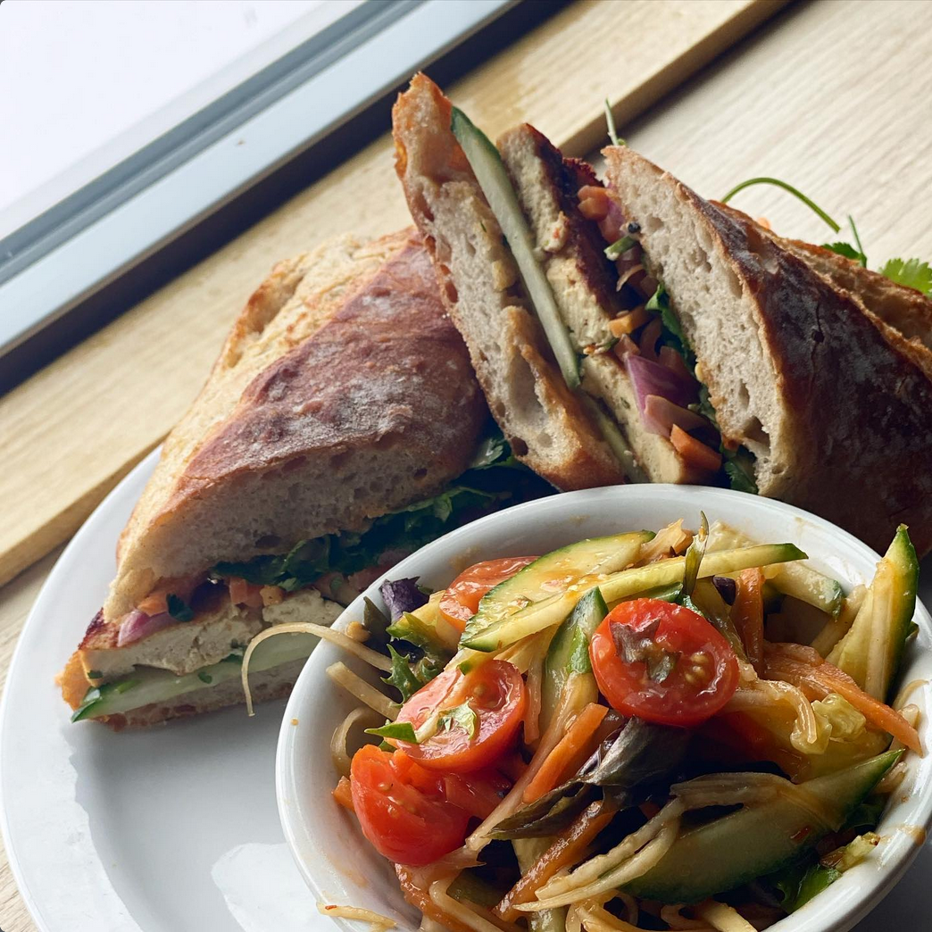 Sandwich and salad on a plate by a window. The sandwich is on crusty bread, filled with vegetables and protein. The salad is in a white bowl.