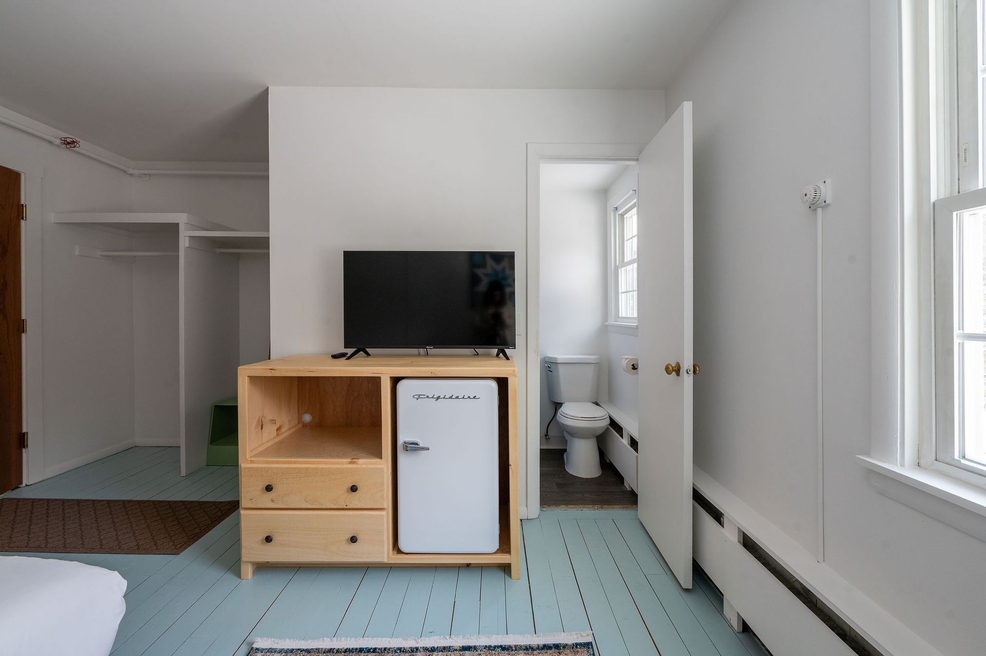 Bedroom with a TV, small refrigerator, and open bathroom door, white walls, and blue flooring.