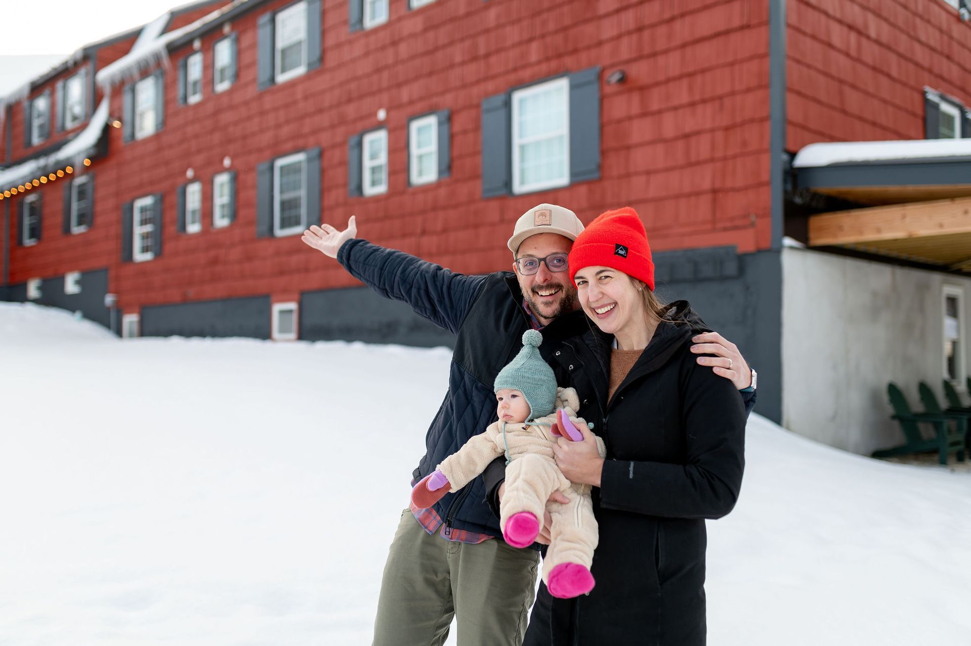 Family poses in snow, red building in background. Man with arm out, woman holds baby.