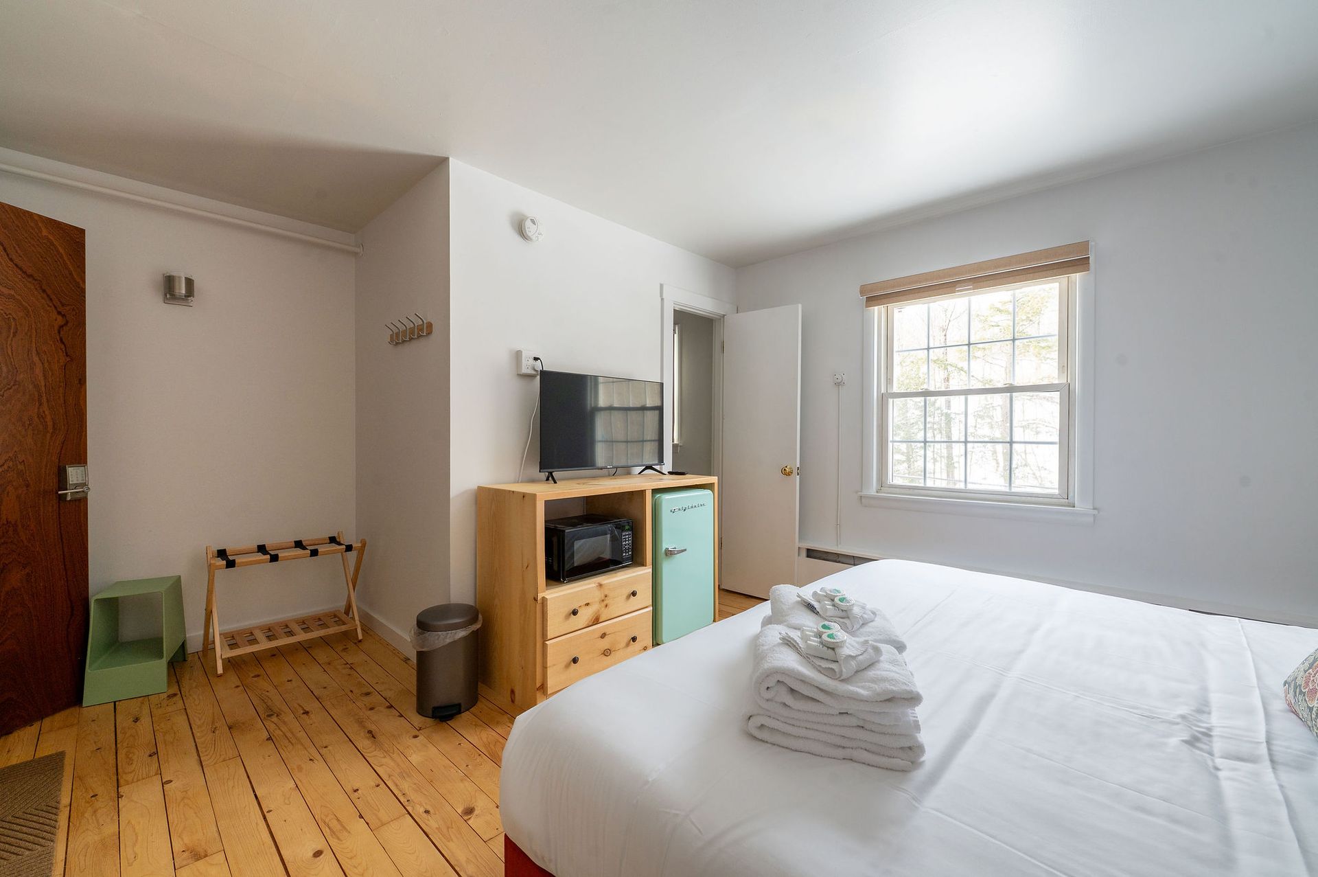 Bedroom with bed, television, window, wooden floors, and a small refrigerator.