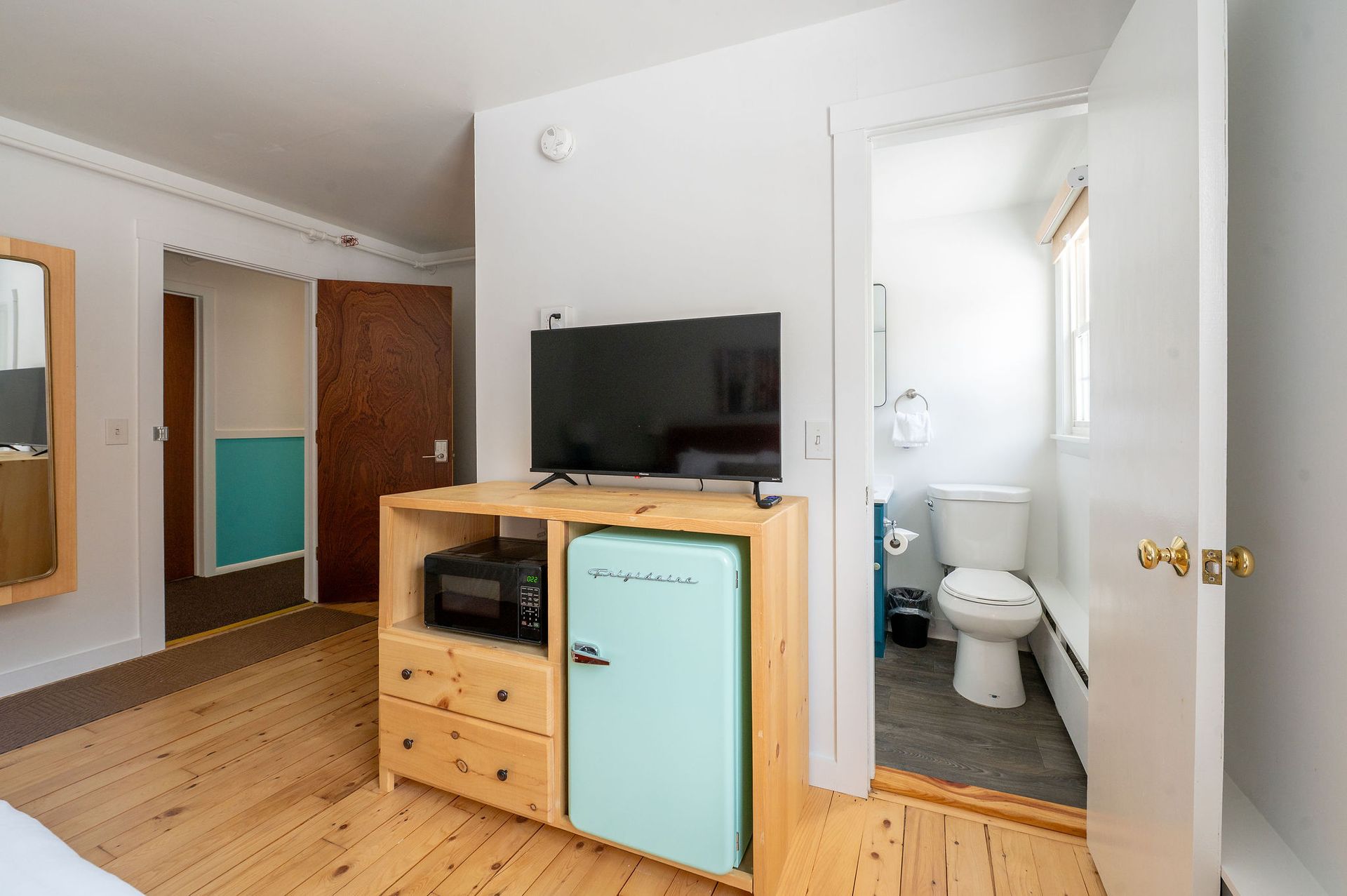 Hotel room with TV, mini-fridge, and bathroom visible. Light wood floors, white walls.