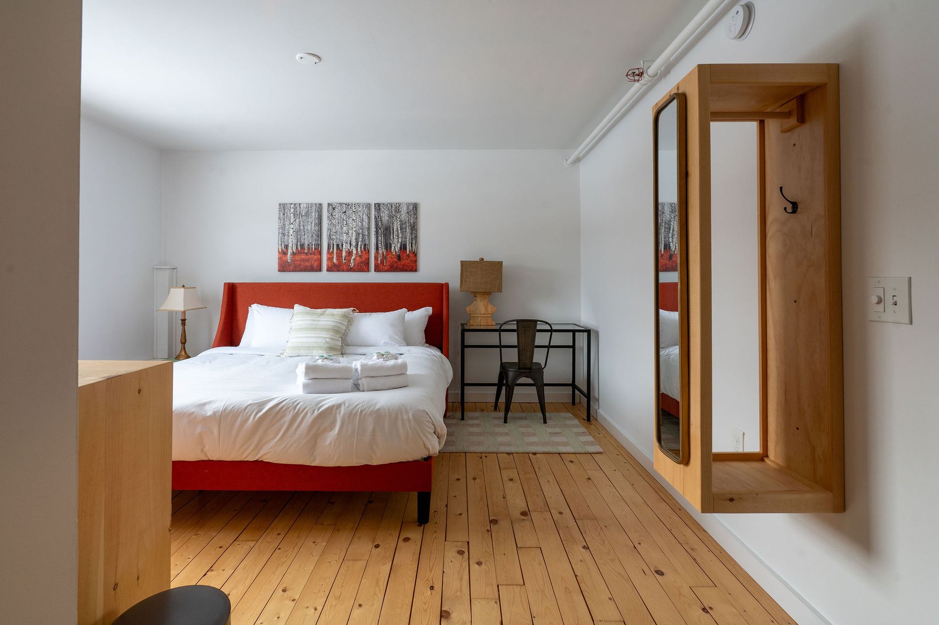 Bedroom with red bed, desk, wooden floor, and wall mirror. Artwork on wall.
