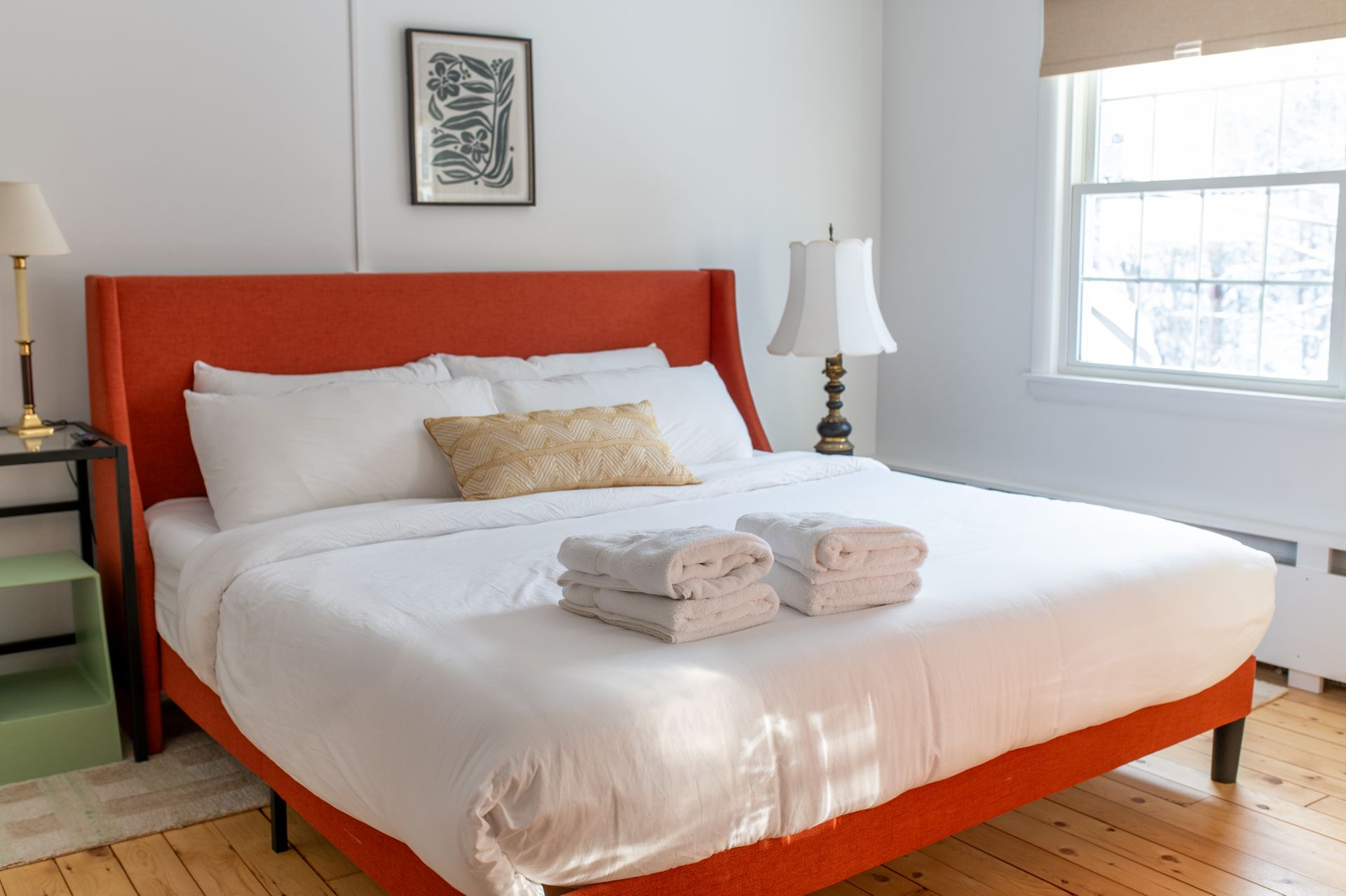 Orange bed with white linens, two stacked towels, two lamps, and a window.