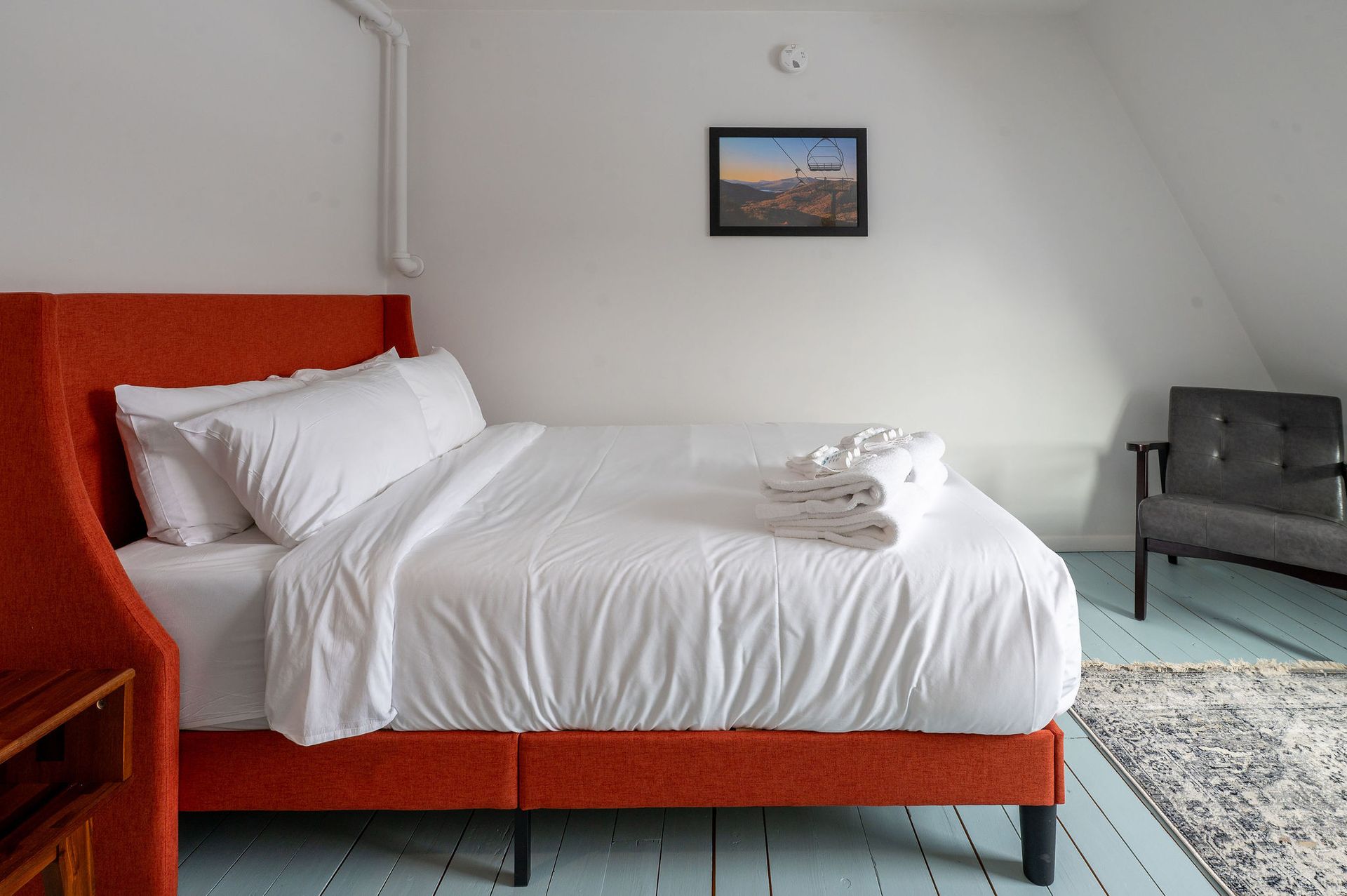 Bedroom with orange bed, white bedding, small framed art, gray chair, and patterned rug.
