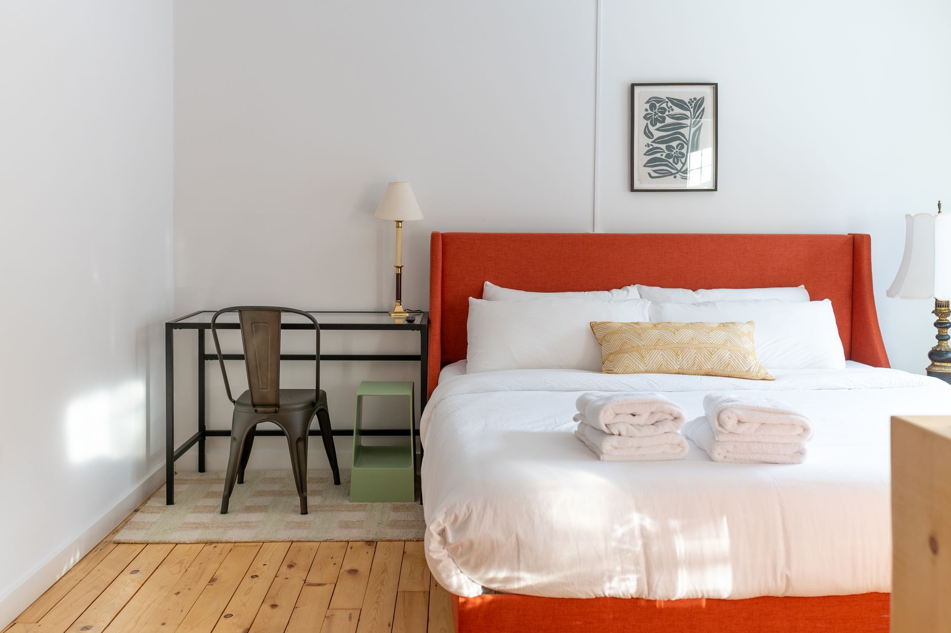 Bedroom with orange headboard, desk, chair, and art on a white wall.