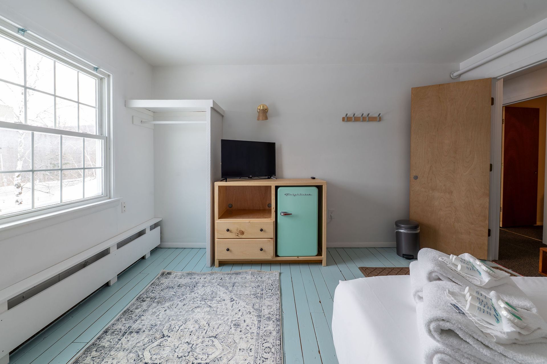 Bedroom with window, closet, small TV, mint fridge, rug, light blue floor, and open door to the right.