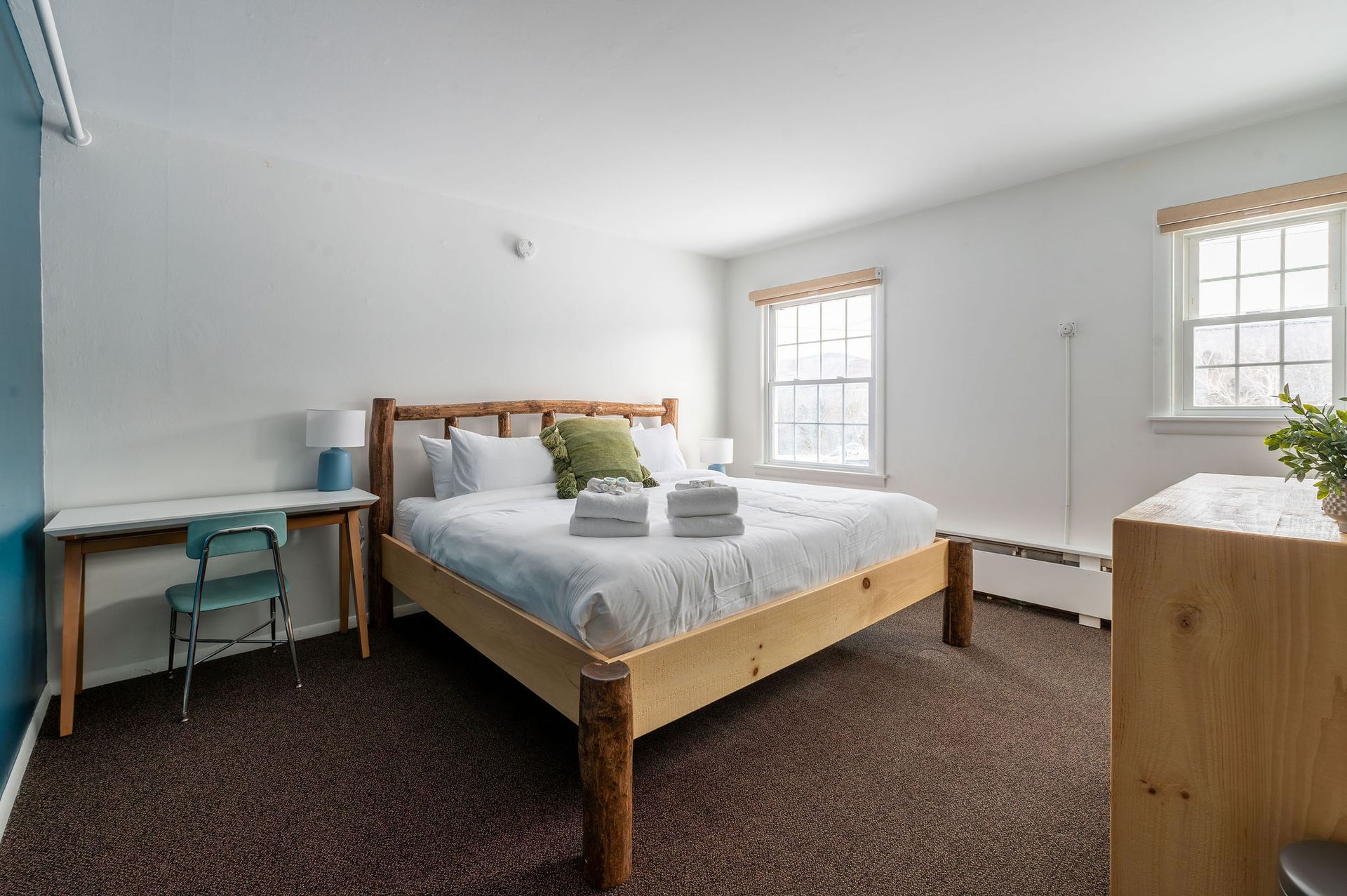 Bedroom with bed, desk, and windows. White walls, wood bed frame, blue accent wall, brown carpet.