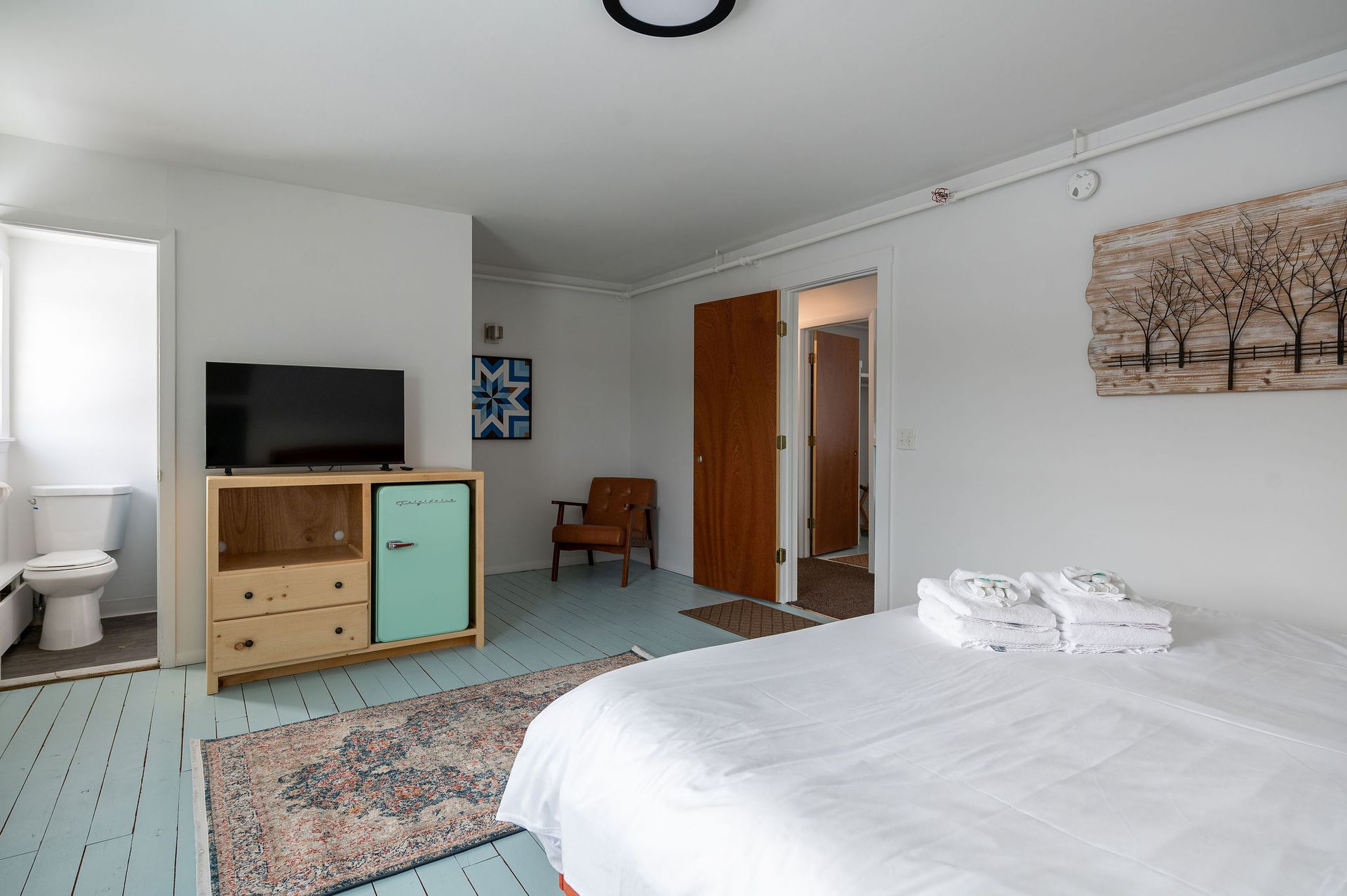 Bedroom with a bed, TV, mini-fridge, and bathroom visible. Blue floor, white walls, wood door.