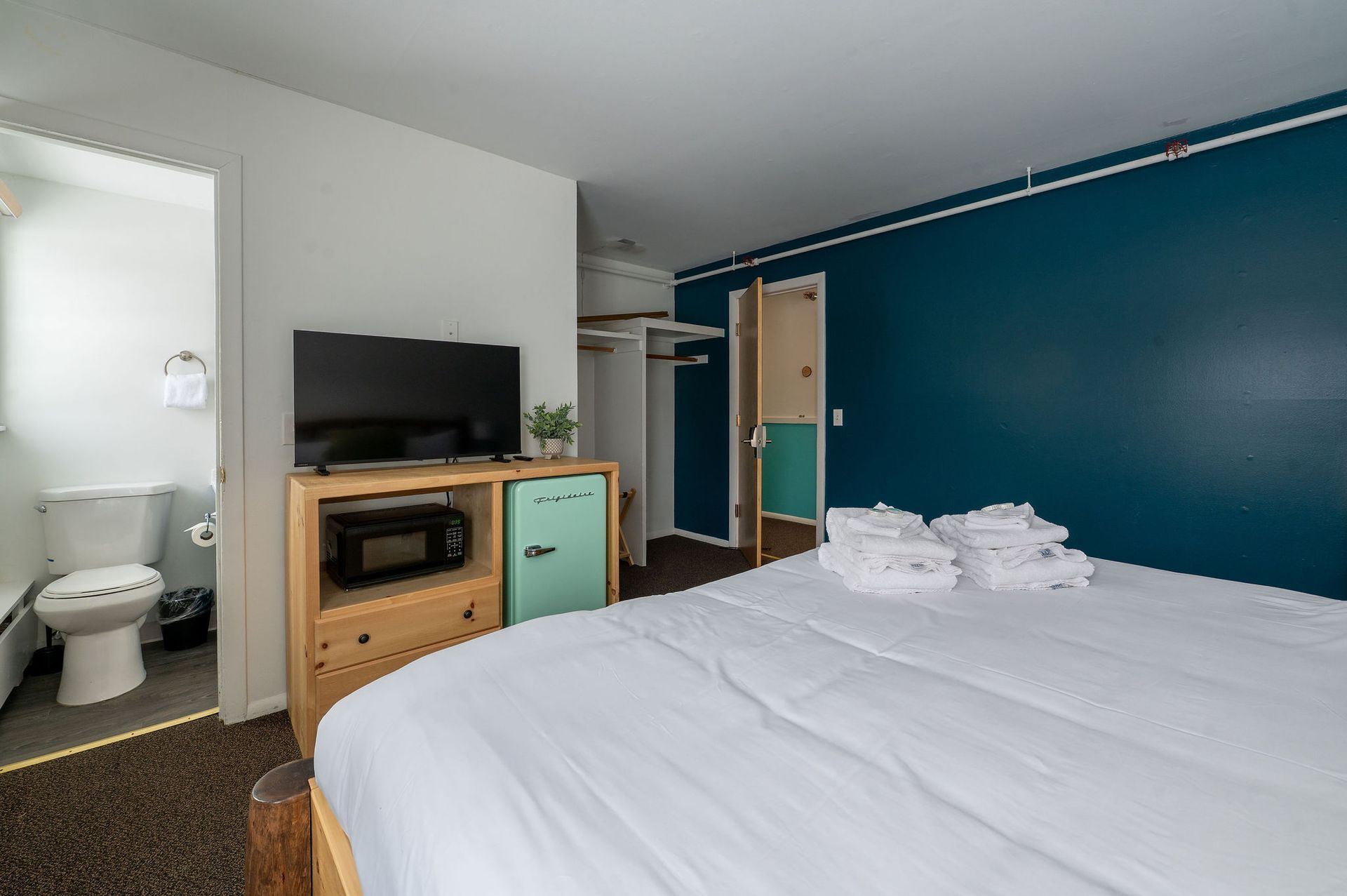 Motel room with bed, TV, microwave, and bathroom visible. Blue accent wall, white bedding, and brown carpet.