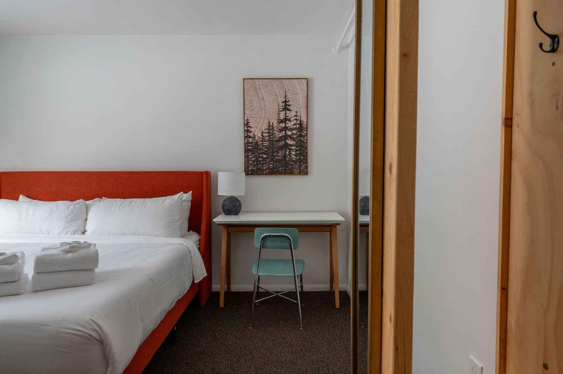 Bedroom with bed, desk, artwork, and mirror. White walls, red headboard, blue chair.