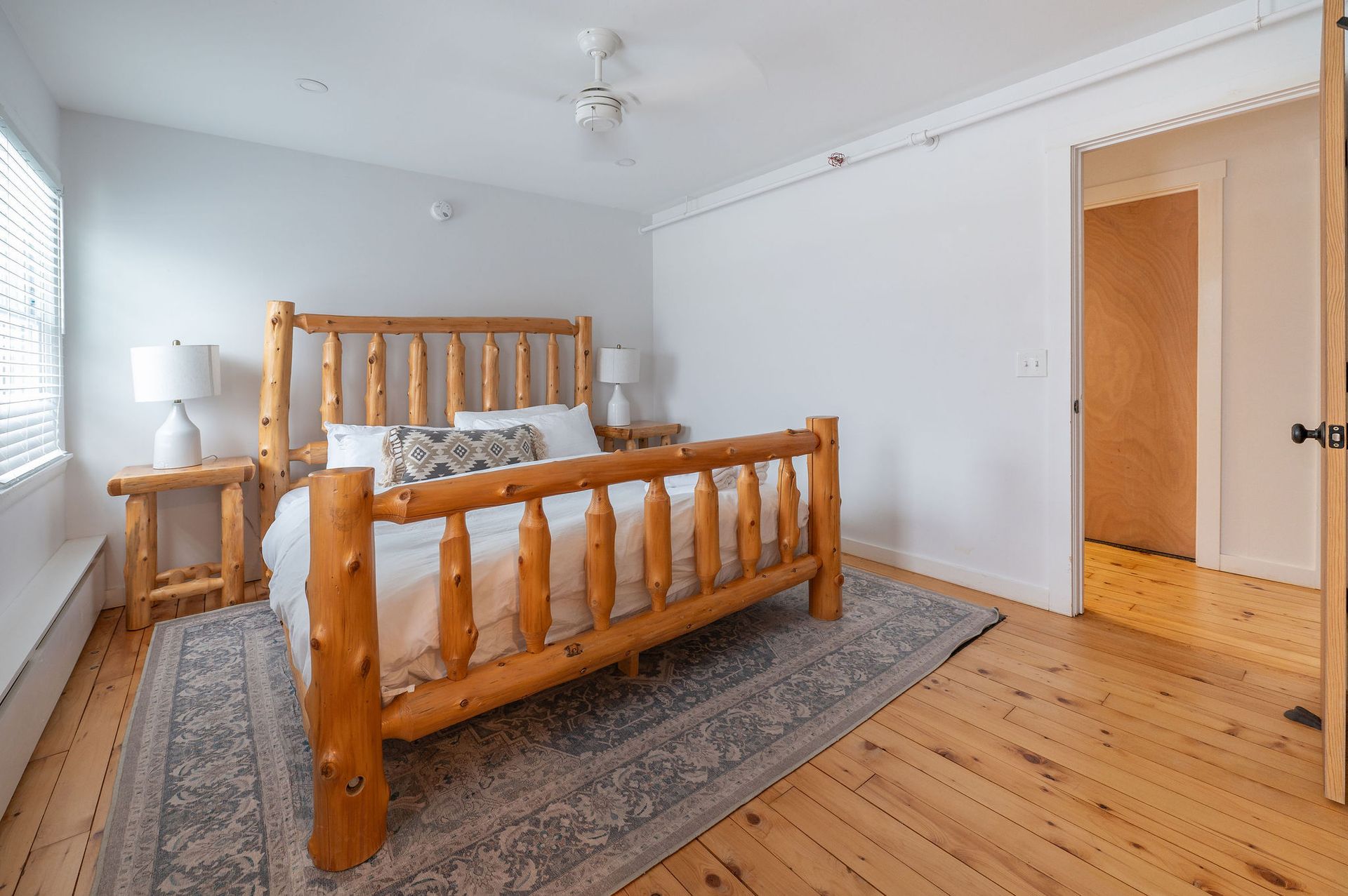 Bedroom with a wooden log bed, white walls, and wood flooring. A rug and lamps are present.