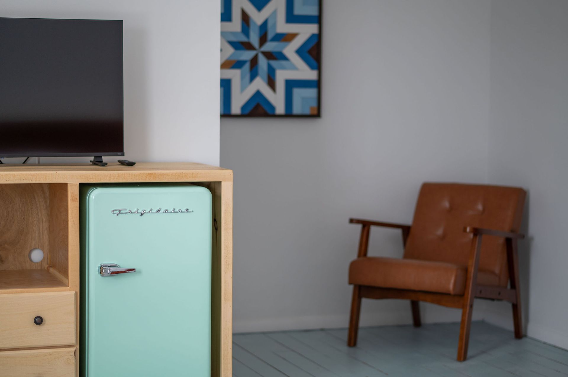 A mint-colored mini-fridge in a wooden cabinet, beside a brown leather armchair. A blue and brown geometric art piece hangs on the wall.