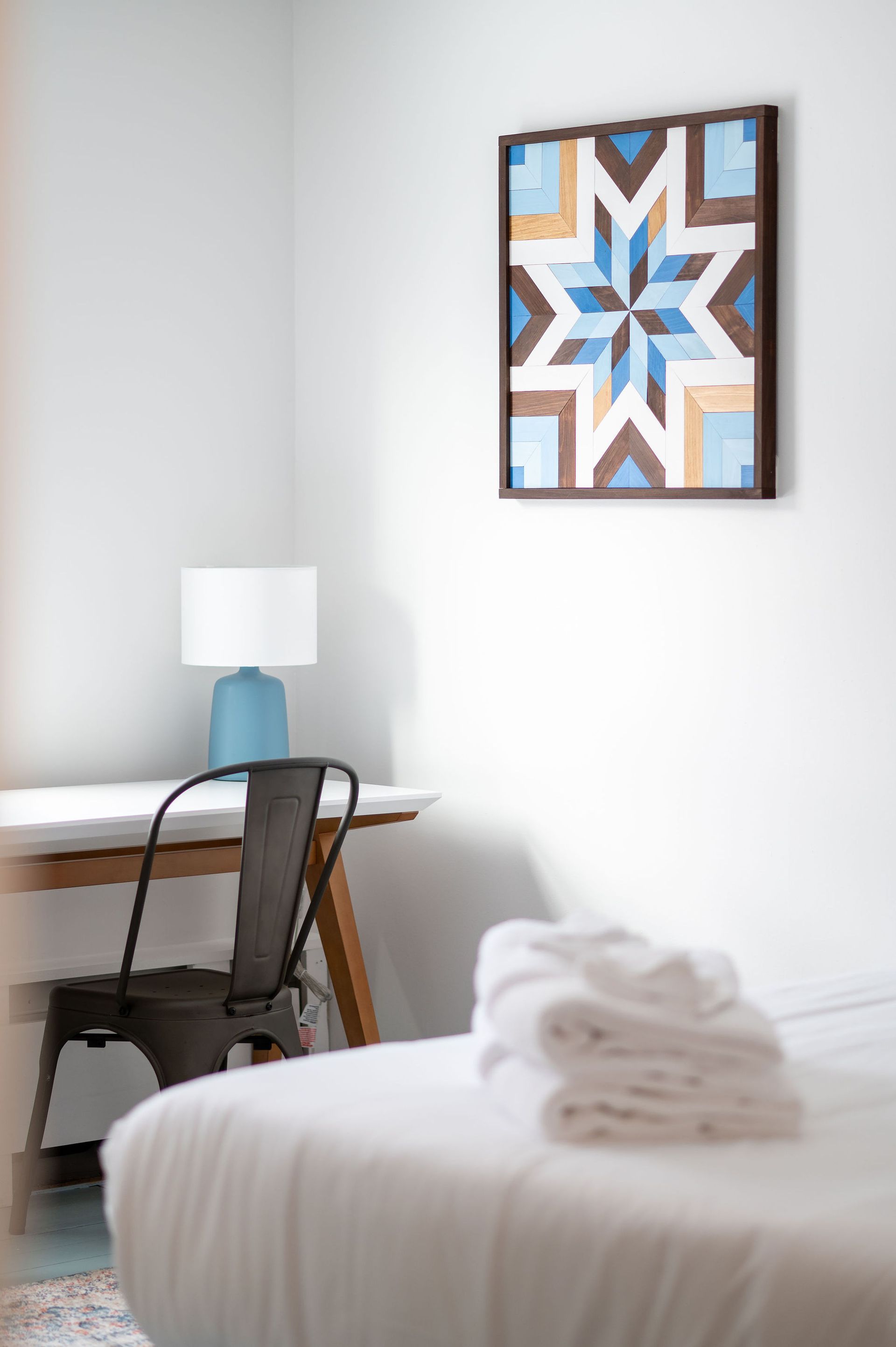 Bedroom corner with desk, chair, and art; white bed with folded towels, blue lamp.