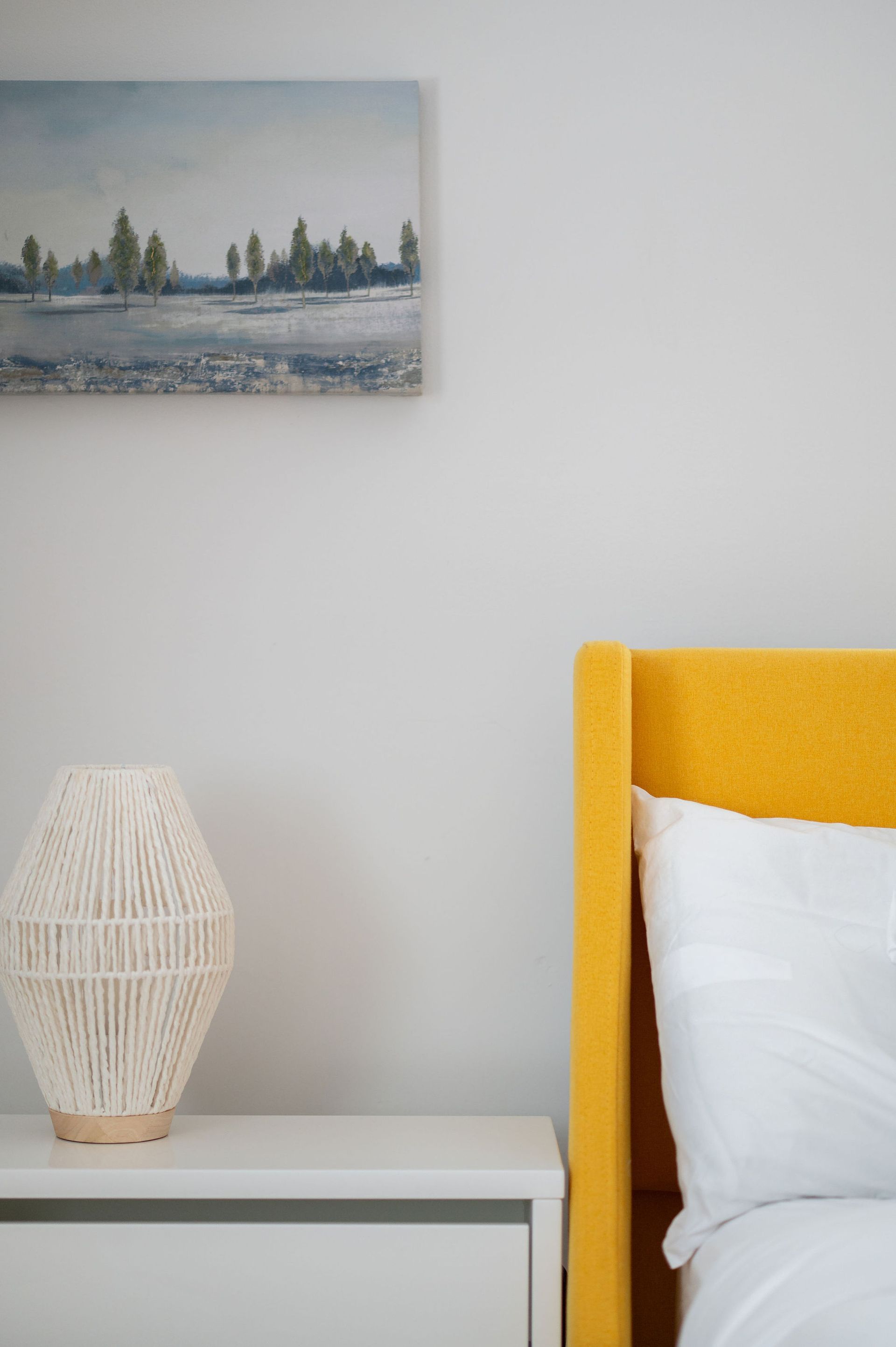 Bedroom with yellow bed frame, white nightstand, lamp, and landscape painting.